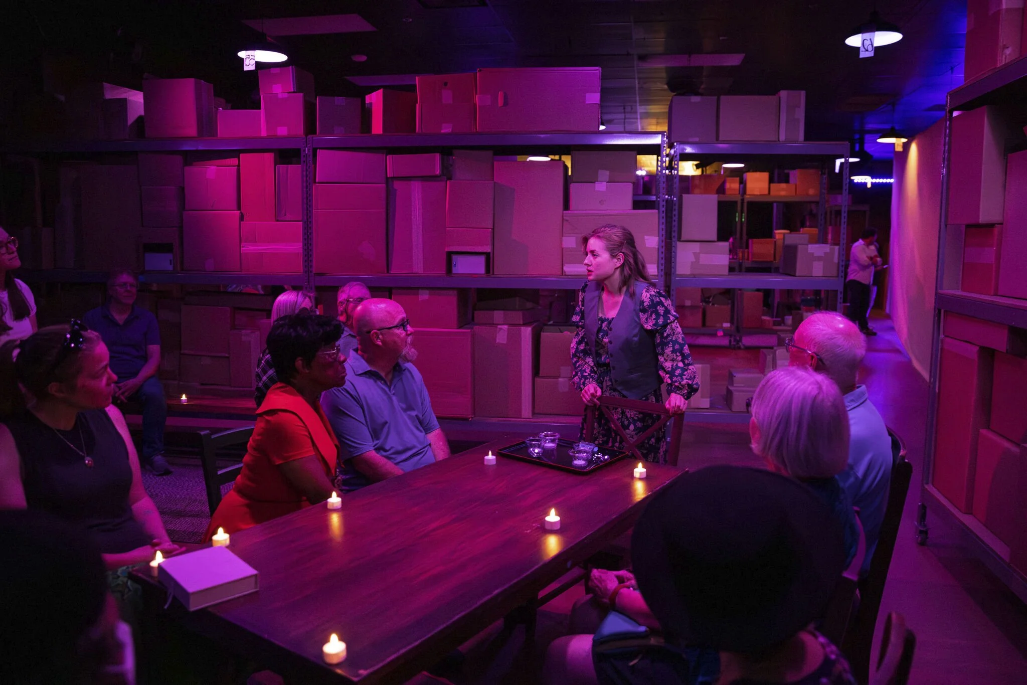 A group of people sitting around a table in a dark room illuminated by purple lighting, with a woman standing and speaking to them, large cardboard boxes on shelves in the background, small candles on the table.