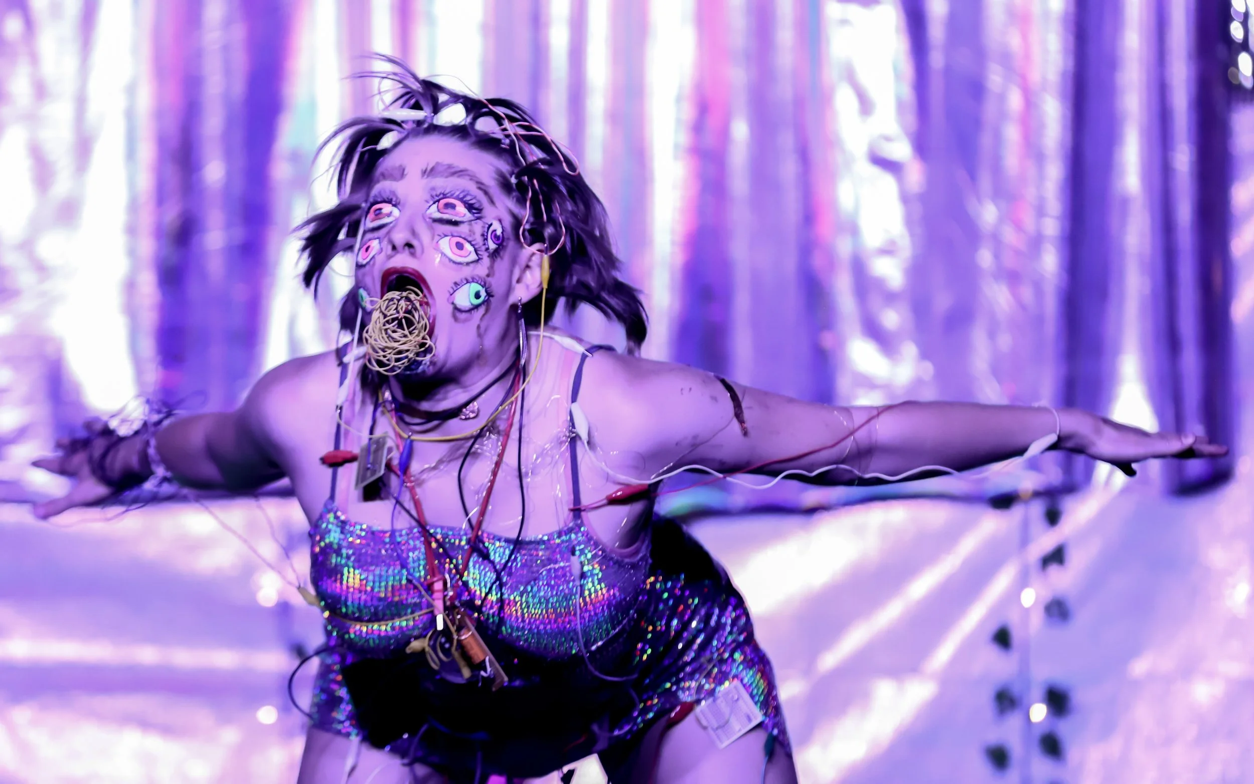 A woman with long, messy hair and multiple eyeball decals on her face is crawling on a shiny, iridescent floor with a purple background. She wears a sparkly, colorful top and has various wires and cables attached to her body.