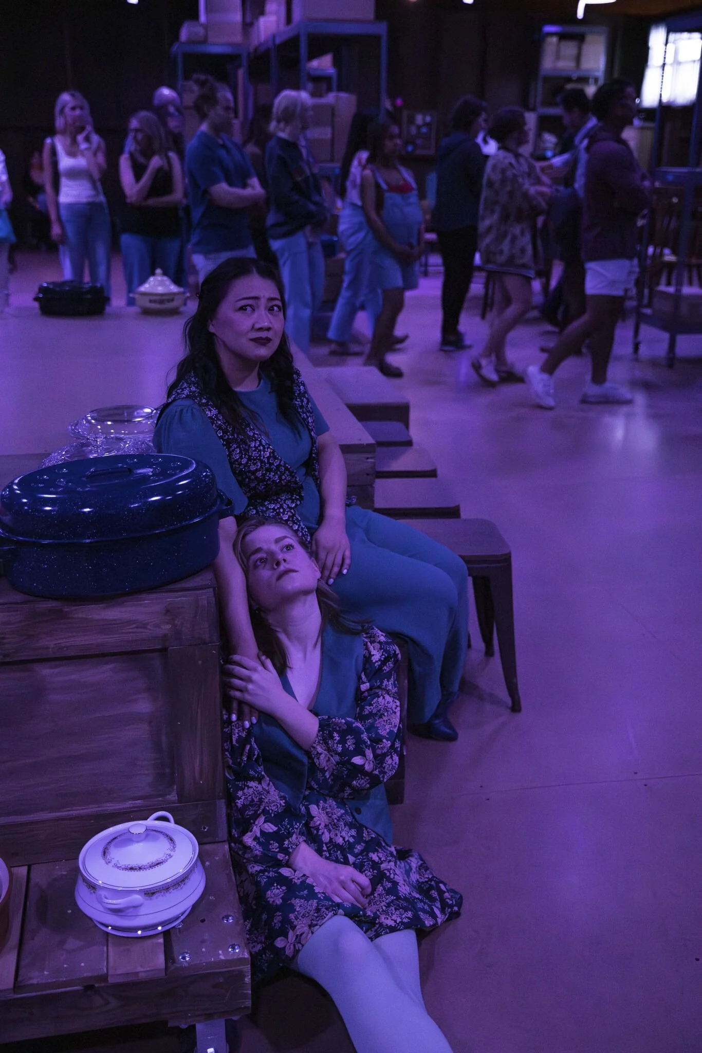 Two women sitting on a bench in a dimly lit room with purple lighting. One woman is lying back with her head on the other's lap, looking upwards. The woman sitting has dark hair and a concerned expression. Behind them, people are queuing or walking, 
