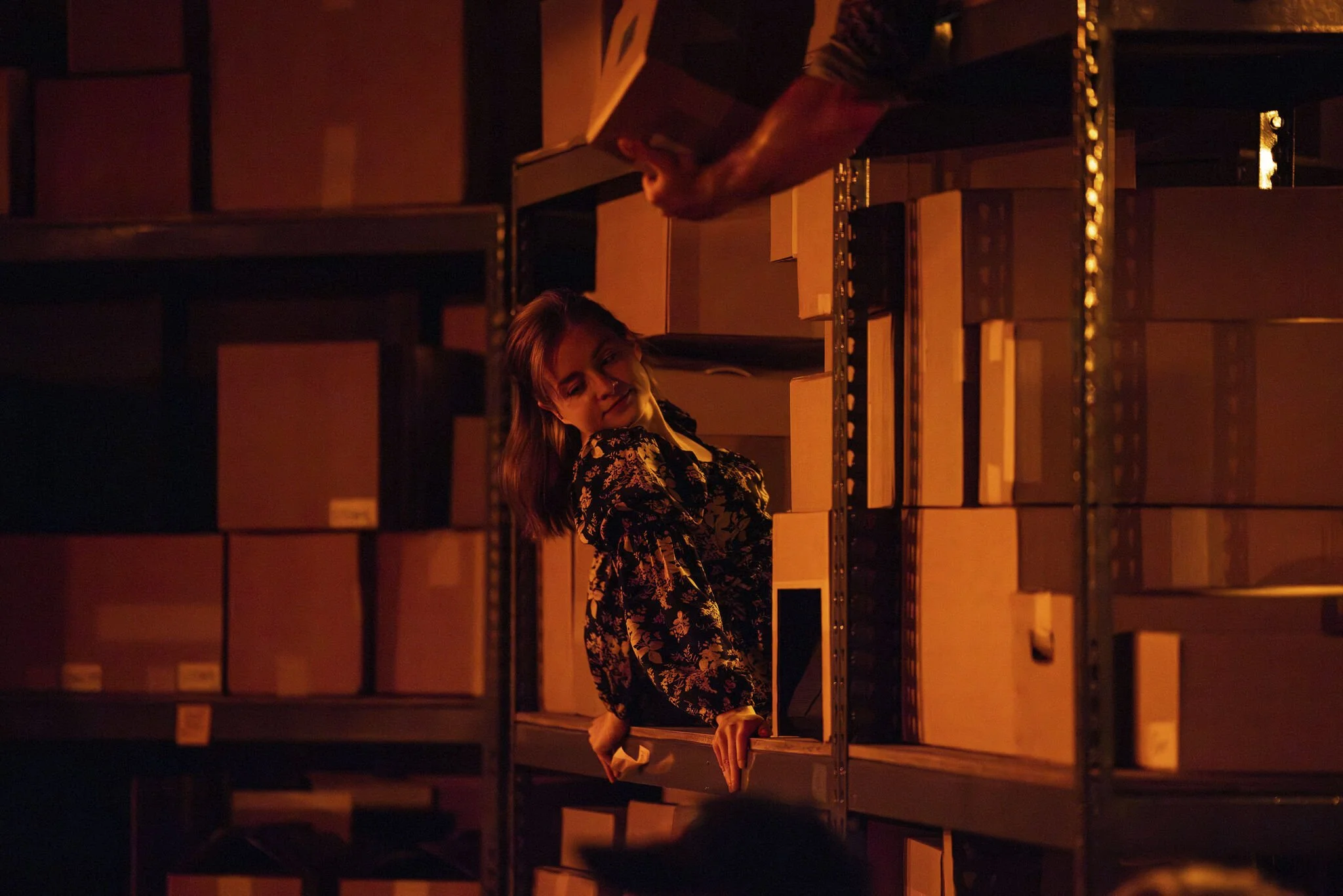 A woman in a floral dress is leaning out of a warehouse shelf filled with cardboard boxes, smiling and looking to her right, with a man in a dark shirt reaching for a box above her.