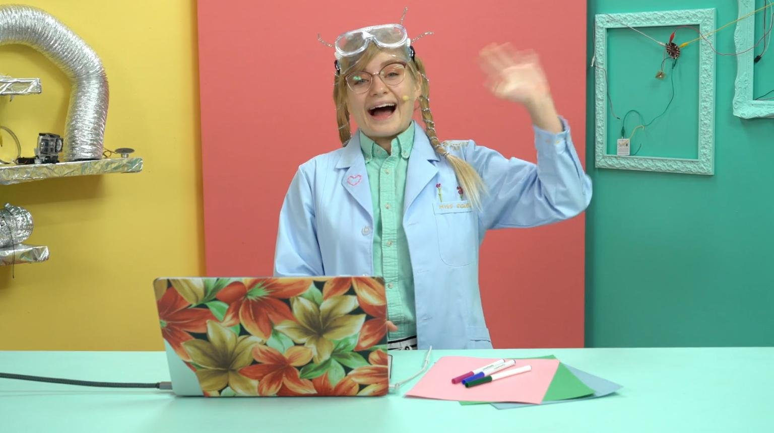 Person wearing safety goggles and a lab coat, waving and smiling in a colorful room with yellow, coral, and teal walls, a table with papers and markers, and shelves with various objects.