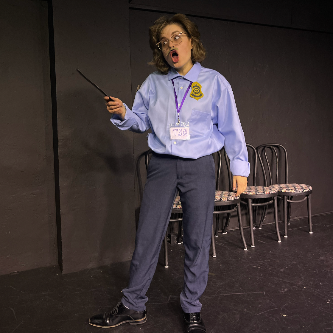 A woman dressed as a security guard, wearing a blue uniform with a badge, glasses, and an ID badge labeled TSA, stands on a stage with a black background and four chairs behind her. She appears to be in the middle of a performance, with her mouth open and holding a black wand.