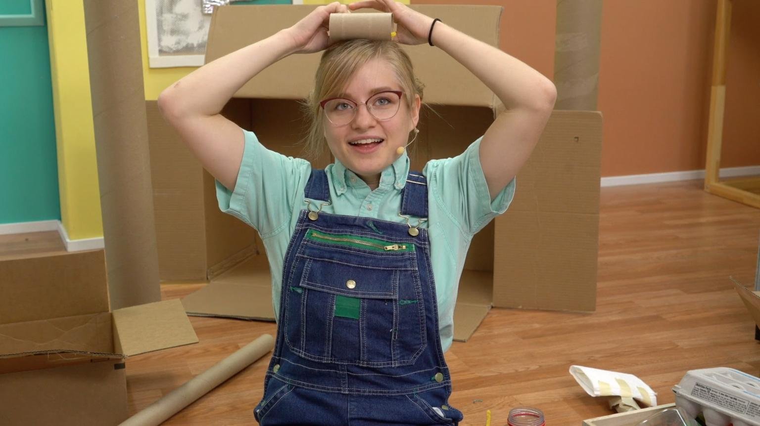 A young woman with glasses and a microphone headset, wearing denim overalls and a mint green shirt, is holding a cardboard tube above her head, smiling, in a room with colorful walls and cardboard structures.