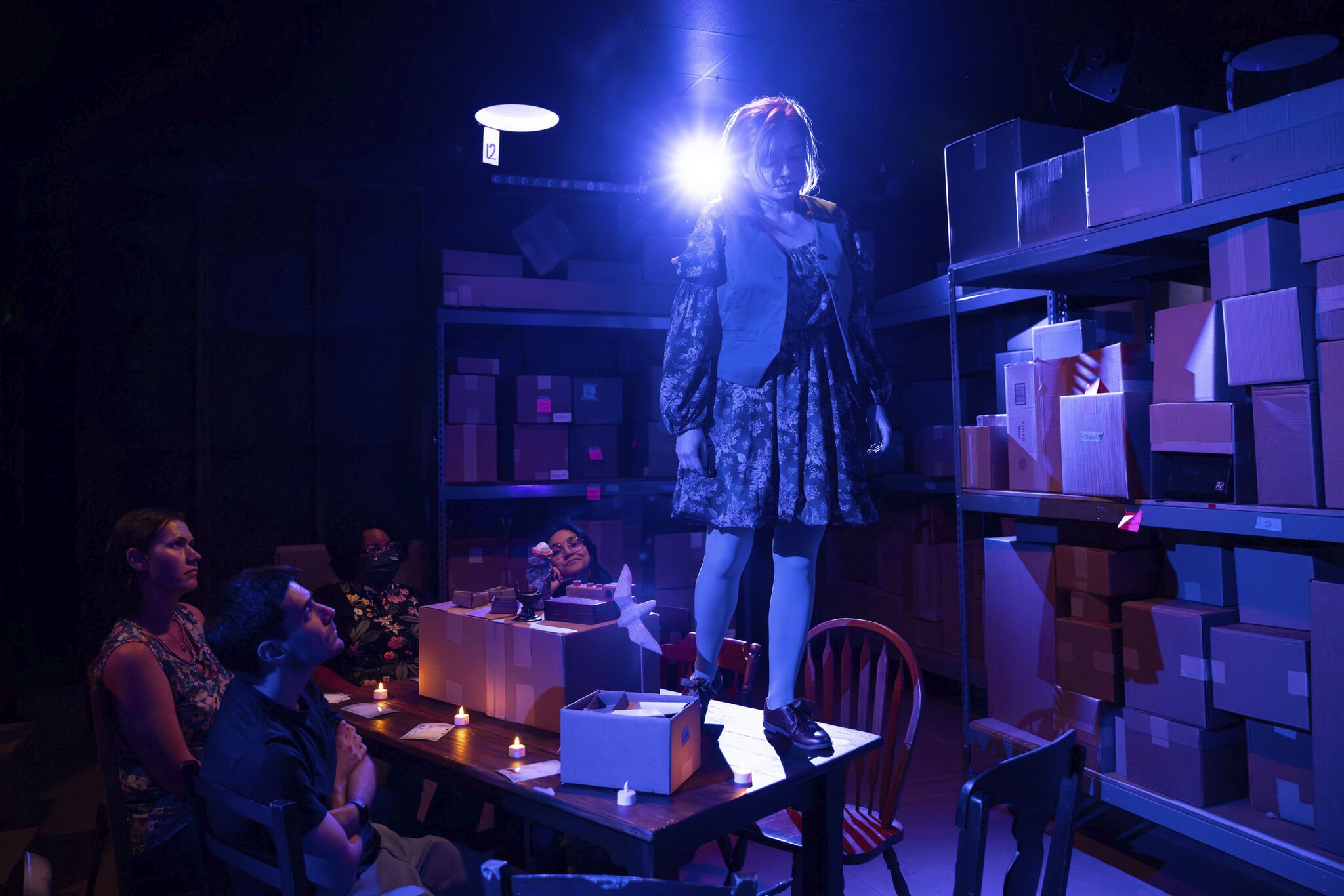 A woman standing on a table in a dimly lit room with purple and blue lighting, surrounded by seated people. The room has shelves filled with boxes and a bright light shining behind her.