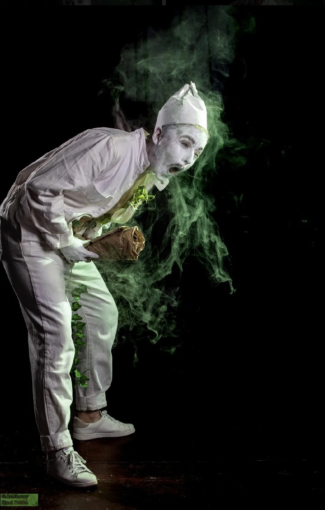 A person dressed as a chef with white makeup or face paint, wearing a chef's hat and white clothing, appears to be gagging or reacting strongly to a smell. There is green smoke or vapor swirling around and behind the person, set against a black backg