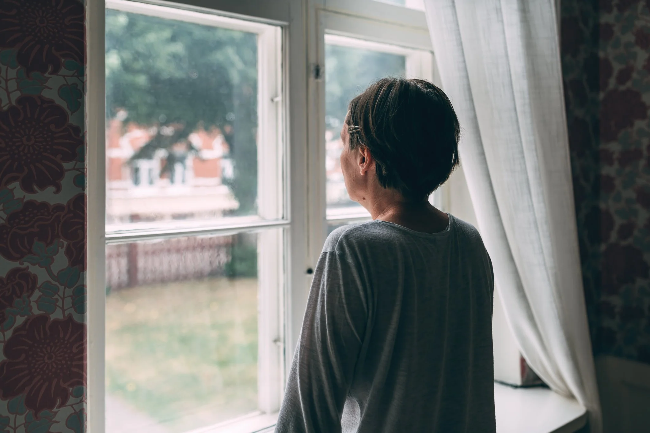 A person with short hair and glasses looking out a window in a room with floral patterned wallpaper and curtains.