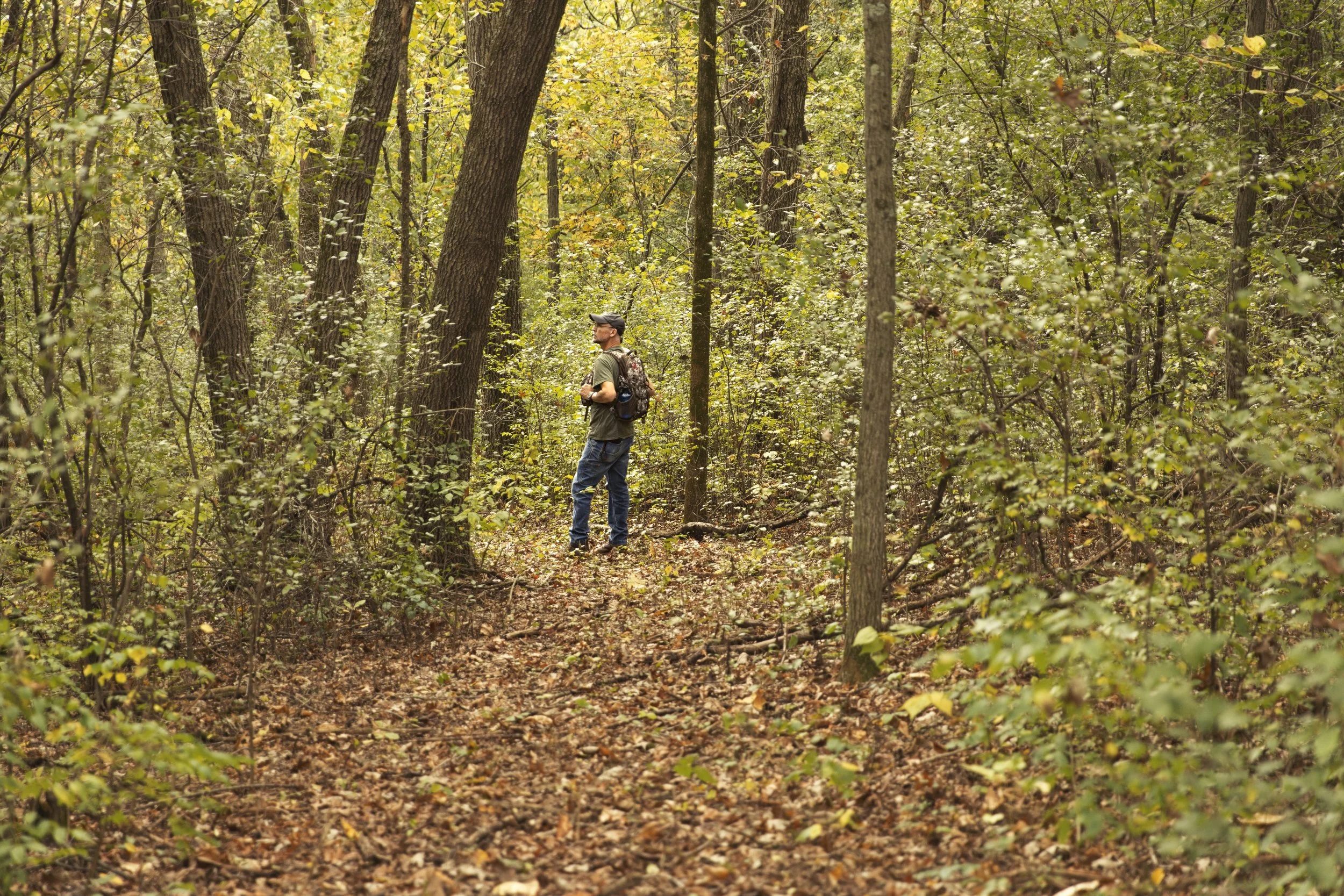 A man stands on a trail in a forest with trees and foliage around him, wearing a cap, glasses, and a backpack.
