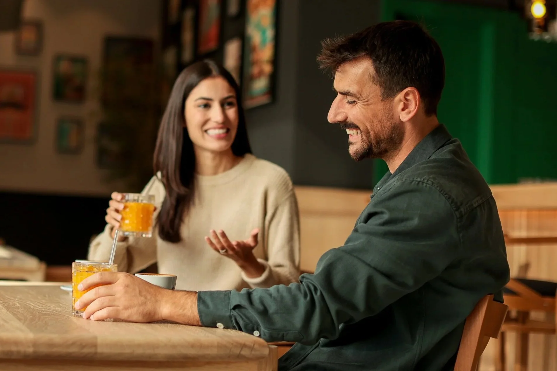 A woman with long dark hair wearing a beige sweater is smiling and holding a glass of orange juice with a straw, engaging in conversation with a man. The man has short dark hair, a beard, and is wearing a green shirt, also smiling. They are sitting at a wooden table in a cozy, dimly-lit restaurant with framed pictures on the wall behind them.