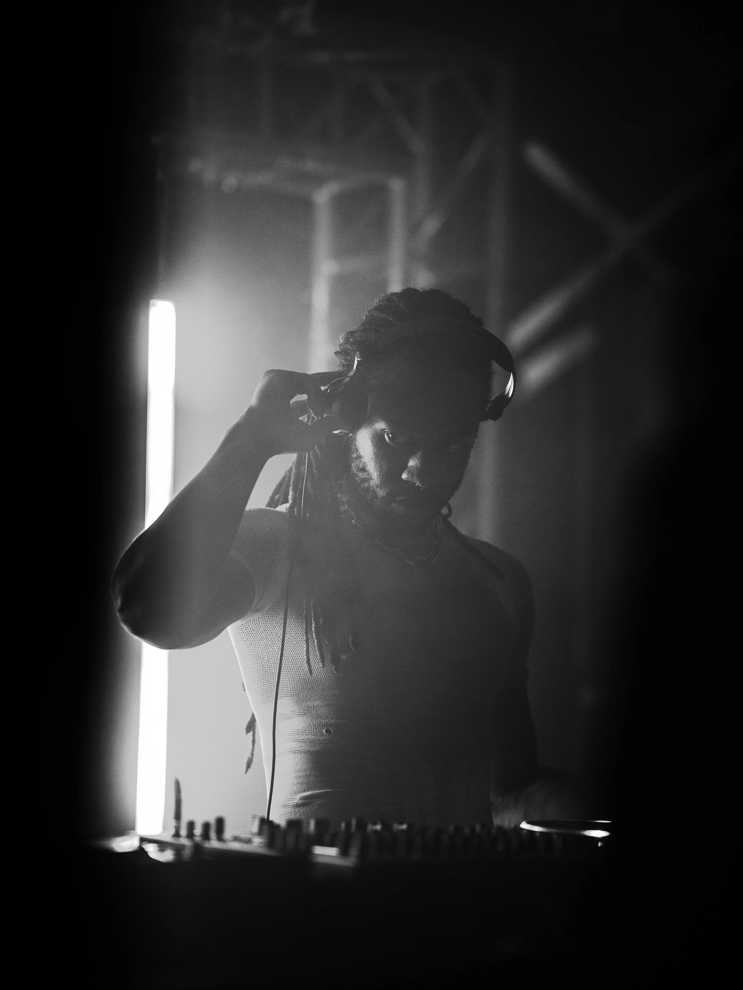 A DJ wearing headphones adjusts his ear while working with DJ equipment in a dark, illuminated setting.