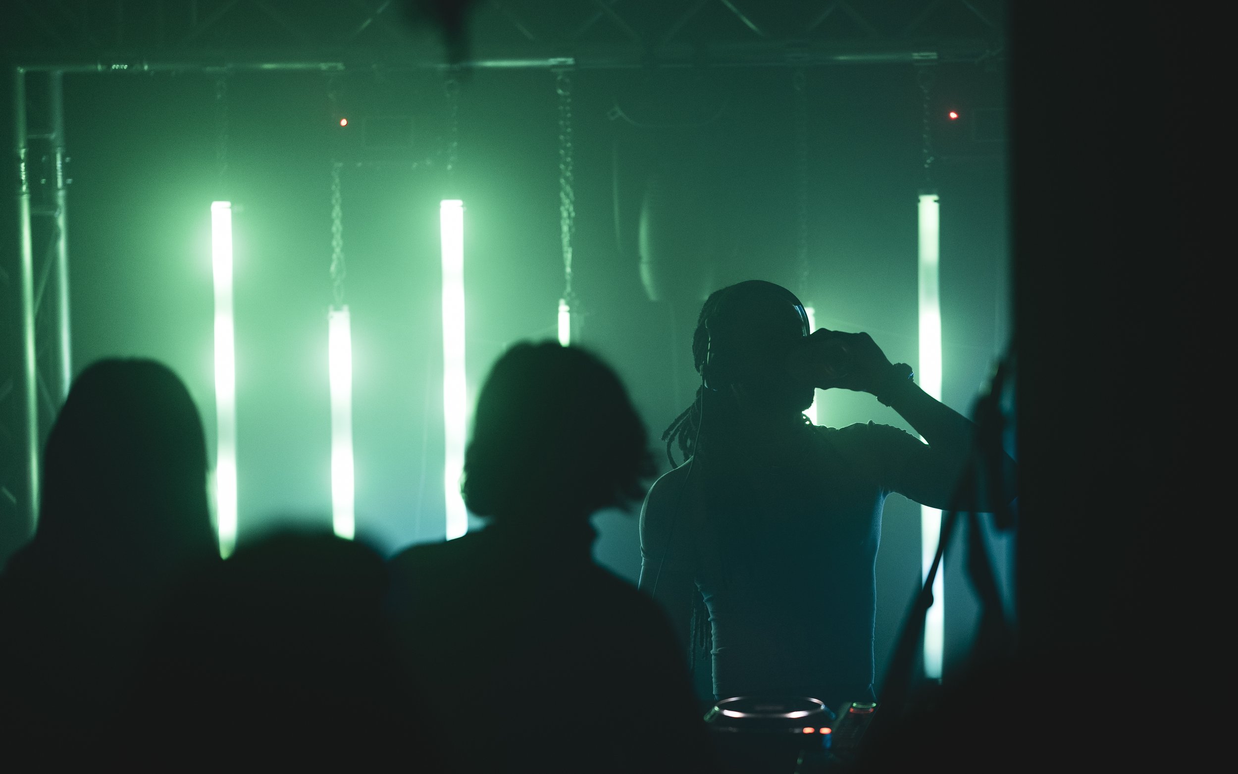 Person wearing headphones and holding a drink at a nightclub with green vertical neon lights in the background.