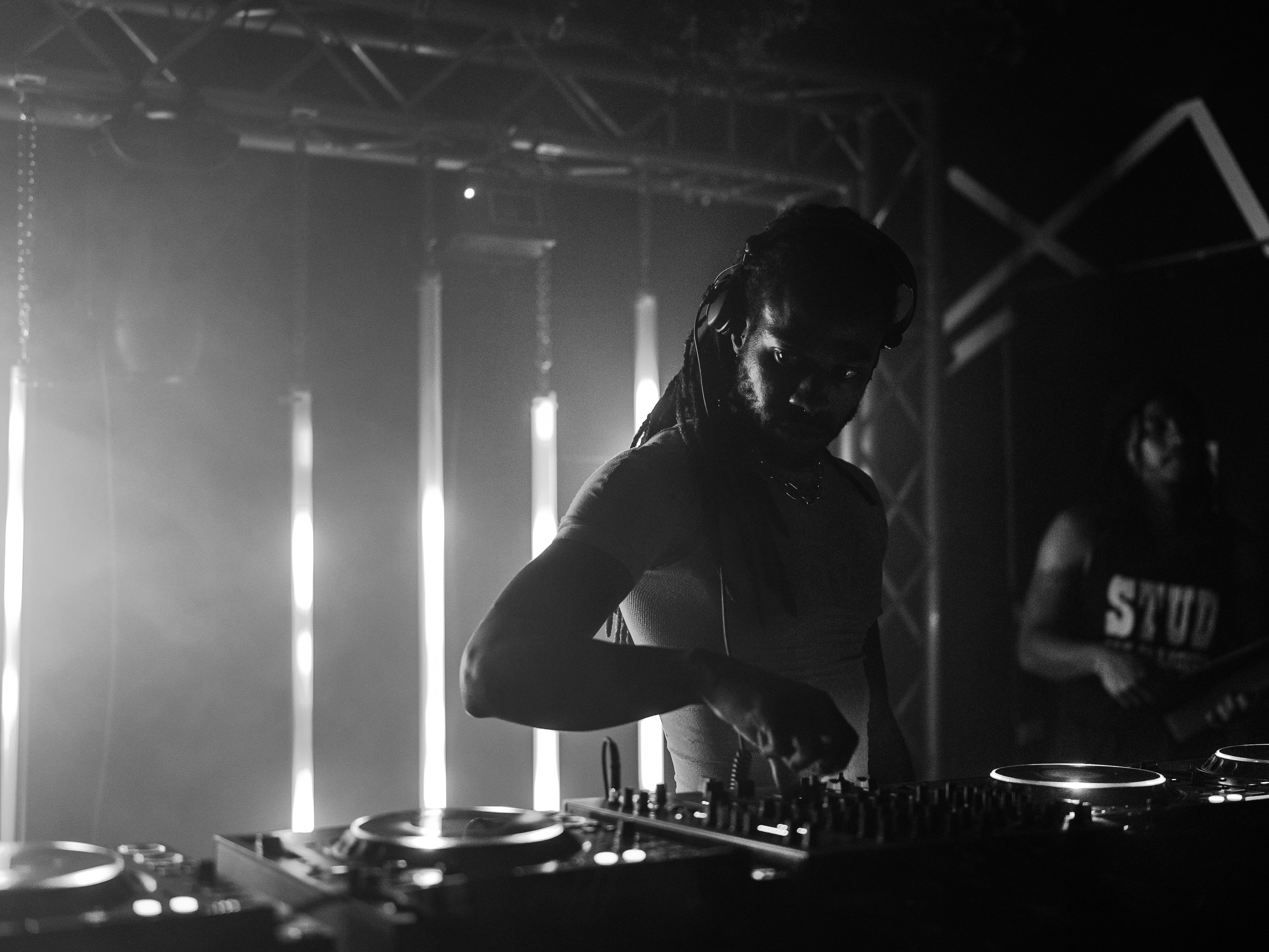 A DJ with dreadlocks wearing a t-shirt and headphones is performing at a dark nightclub, operating a DJ mixer with bright stage lights behind him.