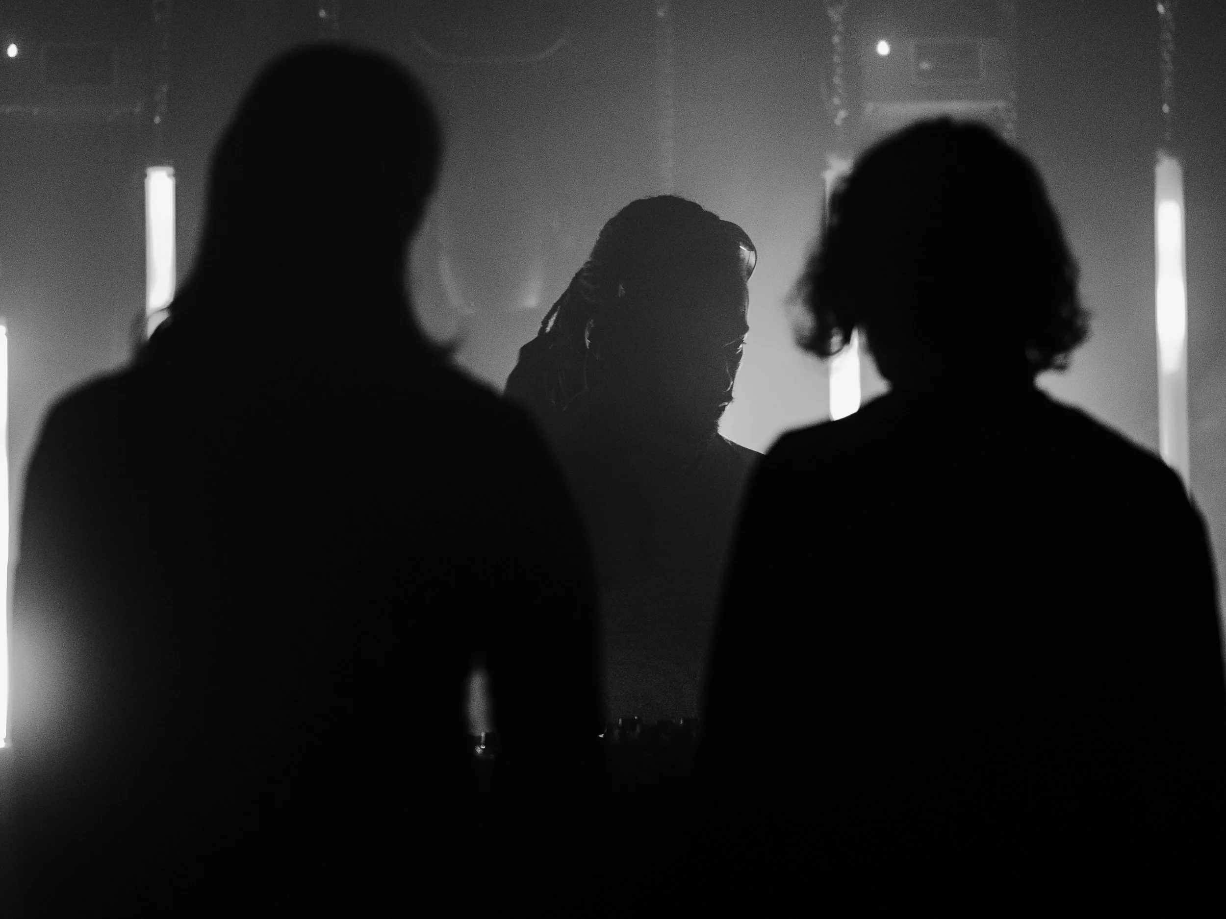 Silhouettes of two people watching a DJ perform on stage with another person in the background, in black and white.