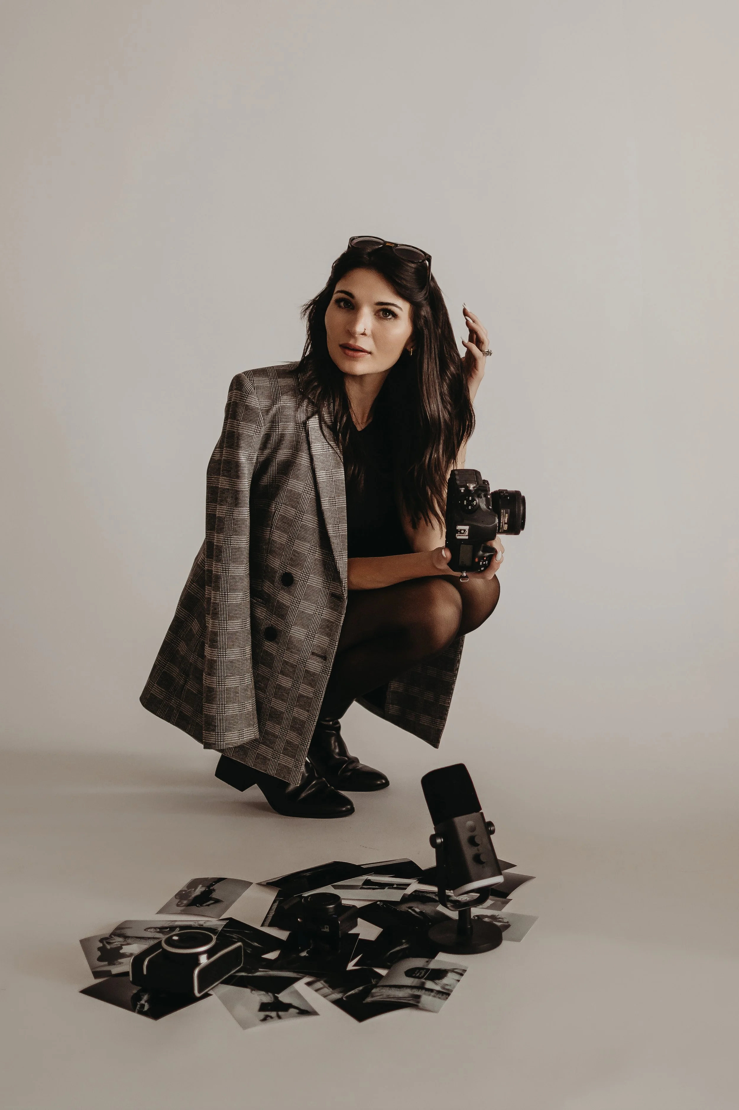 A woman crouching on the floor with a camera in her hand, surrounded by black and white photographs, a microphone, and a camera lens, against a plain white background.