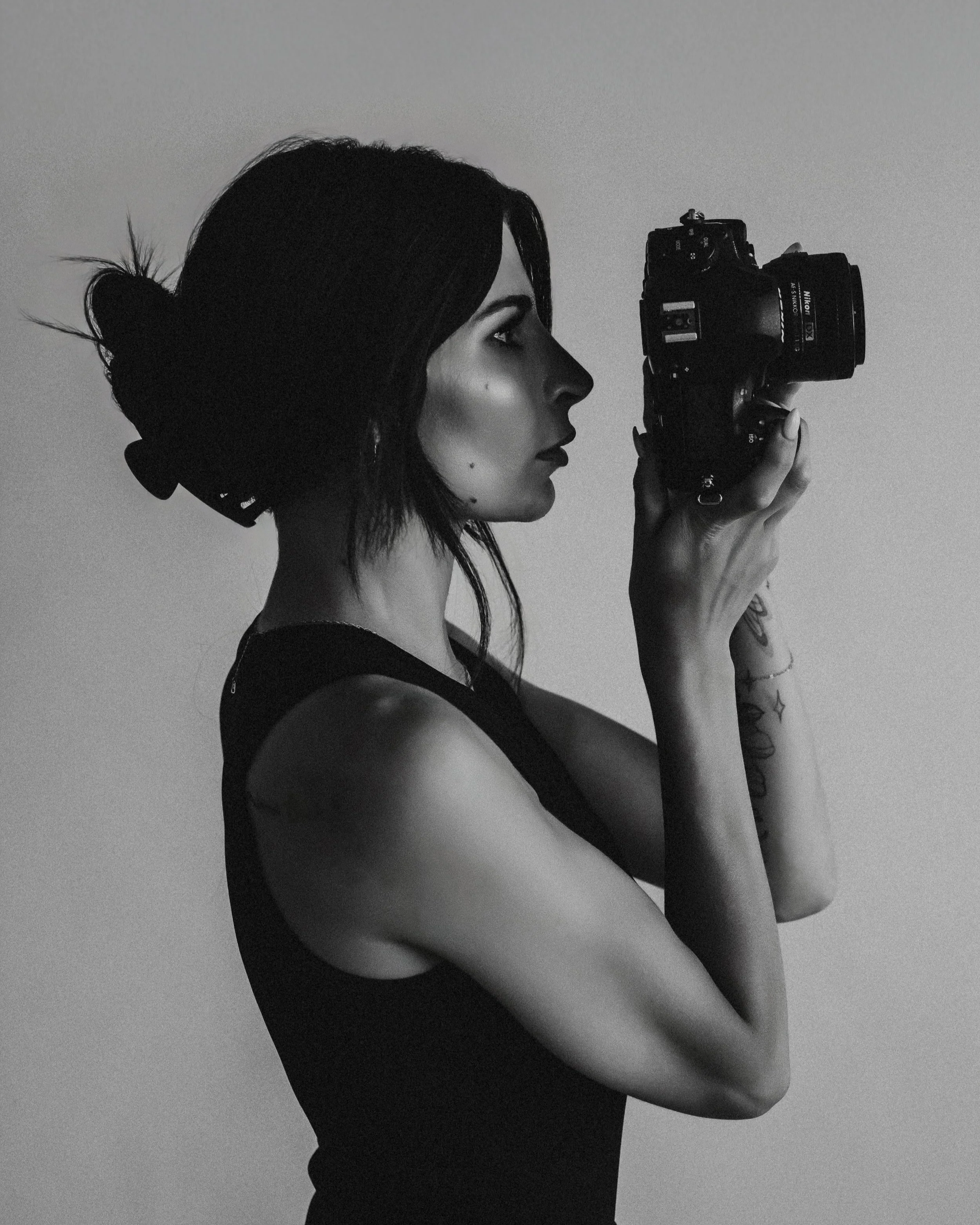 Black and white side profile of a woman with tattoos, holding a camera up to her face.