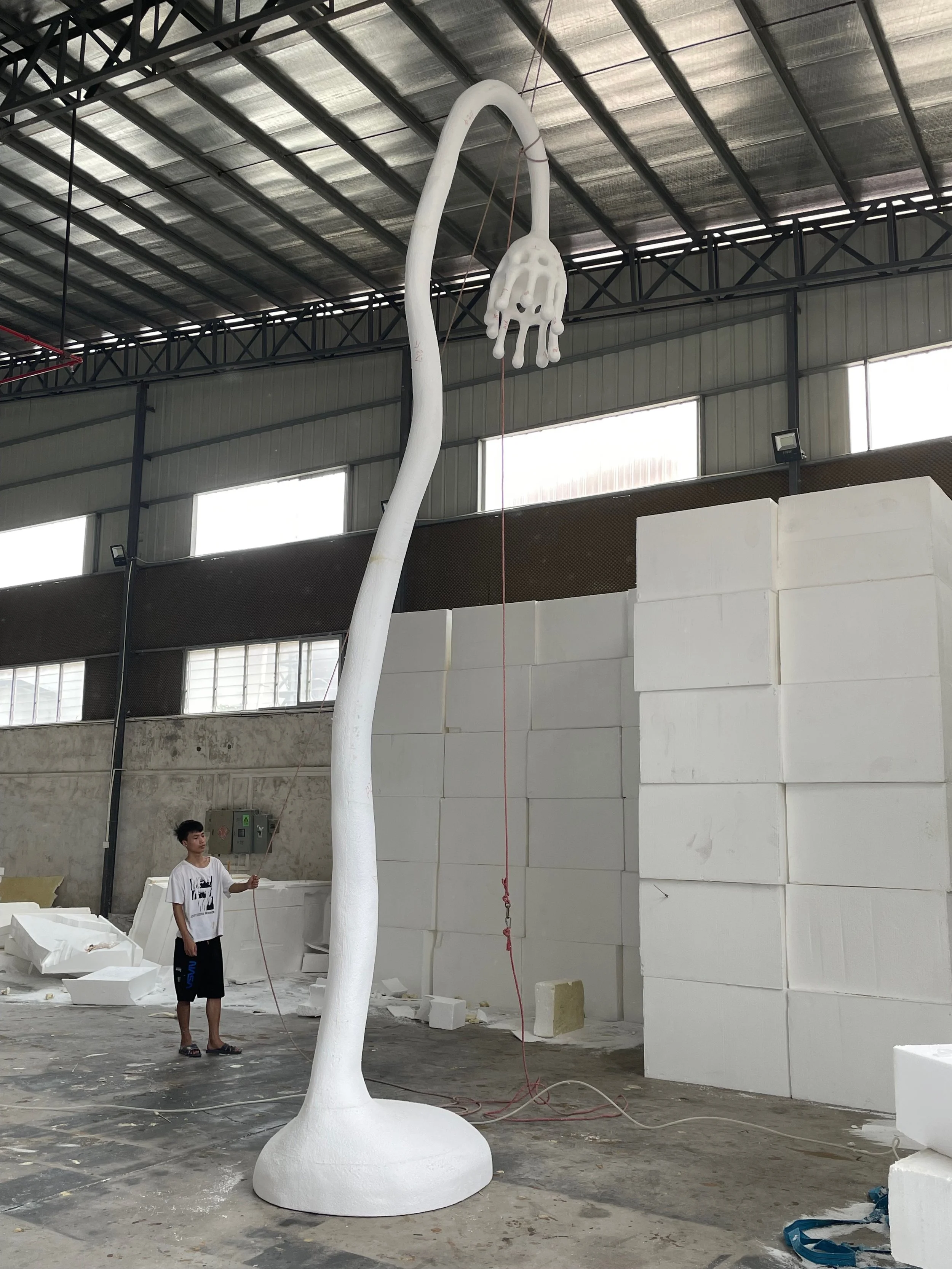 A large, white, abstract sculpture resembling a tall, curved stem with a hand-shaped top in a warehouse with a person standing nearby.