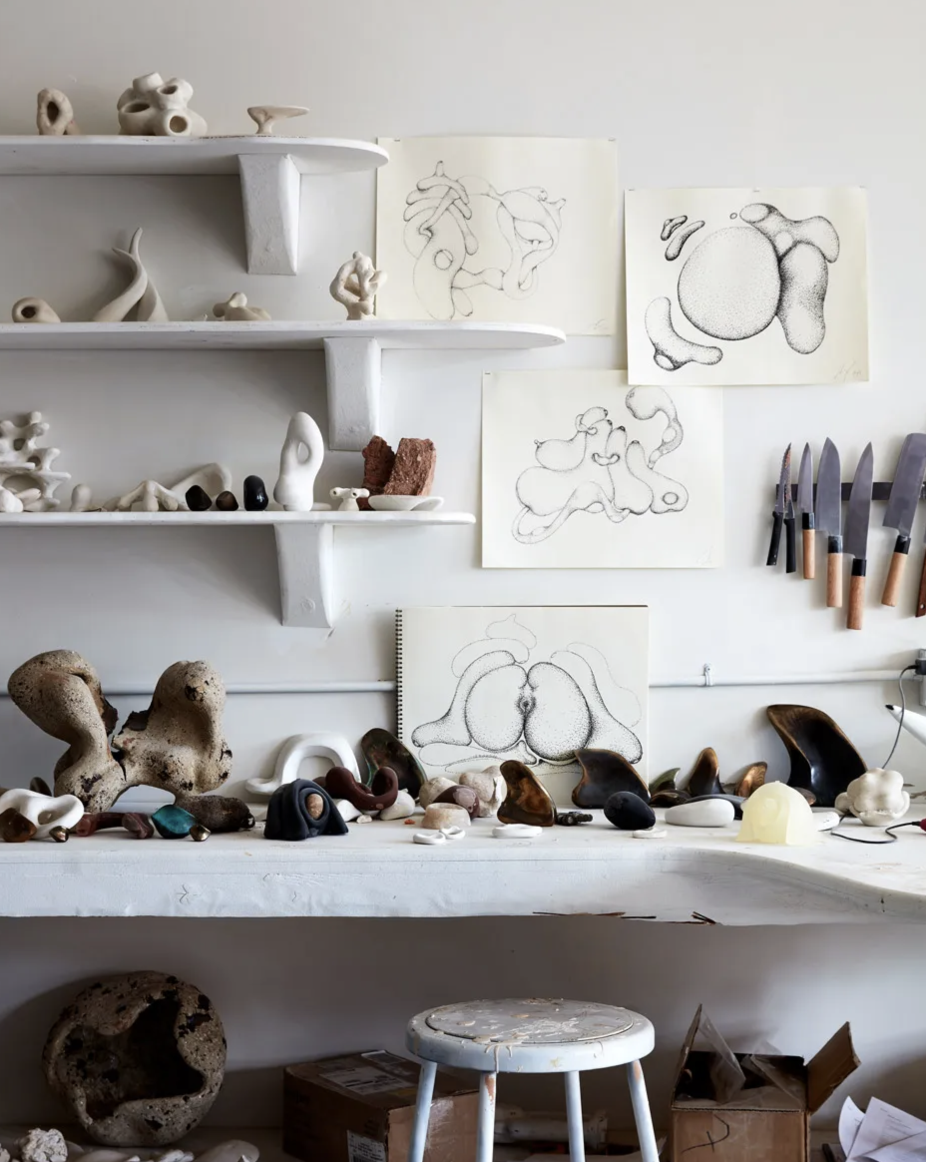Art studio with abstract sculptures, sketches, and tools; shelves and table displaying various pieces and drawings on wall.