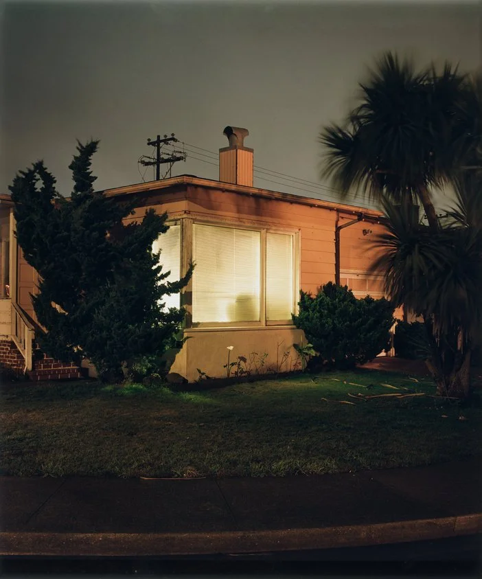 Exterior of a house at night with illuminated window, surrounded by trees and grass.