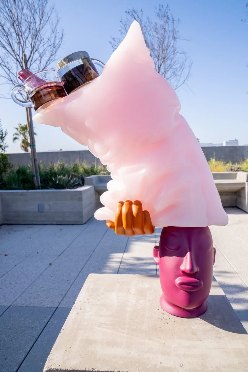 Colorful sculpture of a stylized human head with eyes closed, topped with a large pink cloud-like shape and two glass teapots.