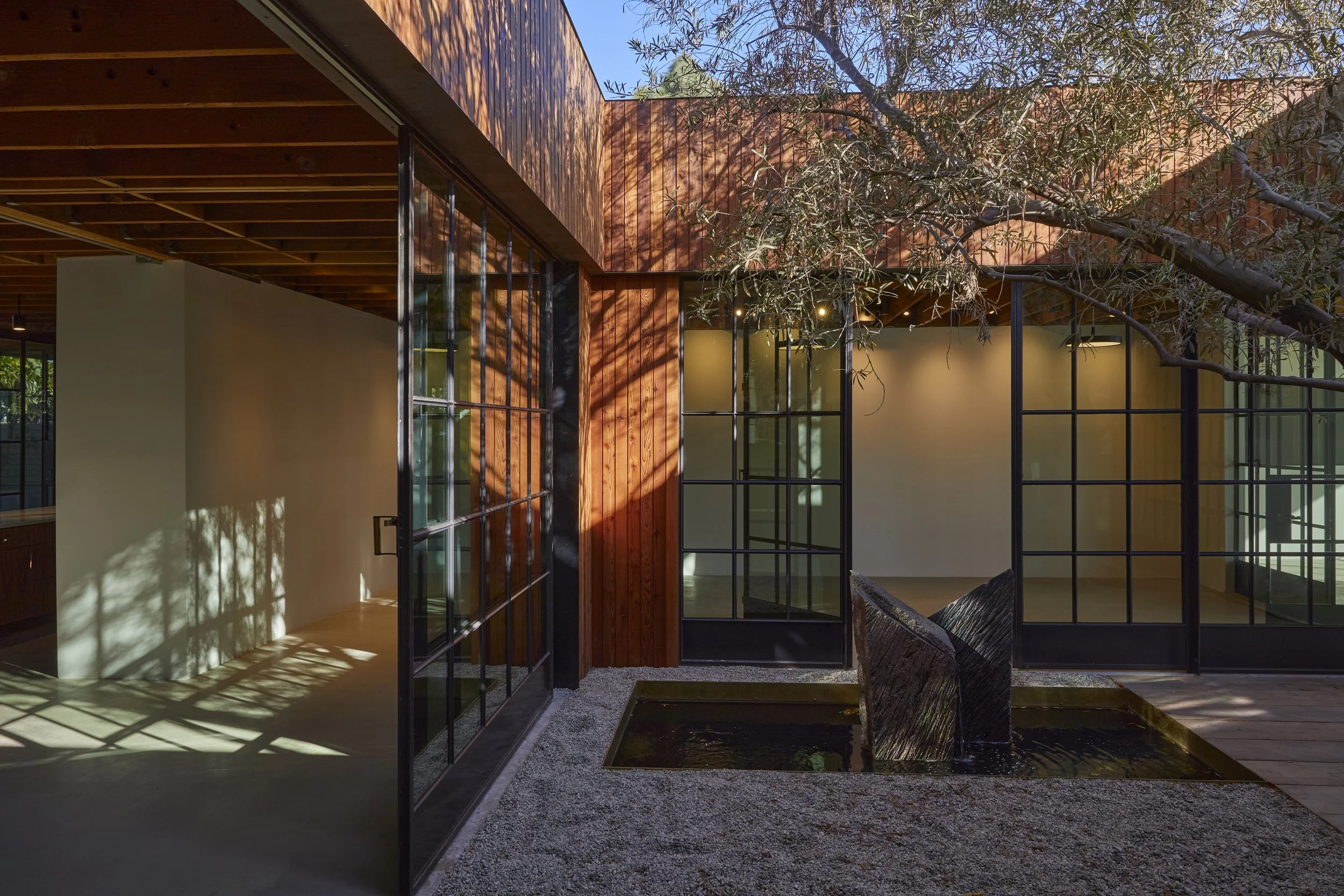 Modern architectural space with glass-paneled walls, wooden ceilings, and a central water feature, surrounded by trees.