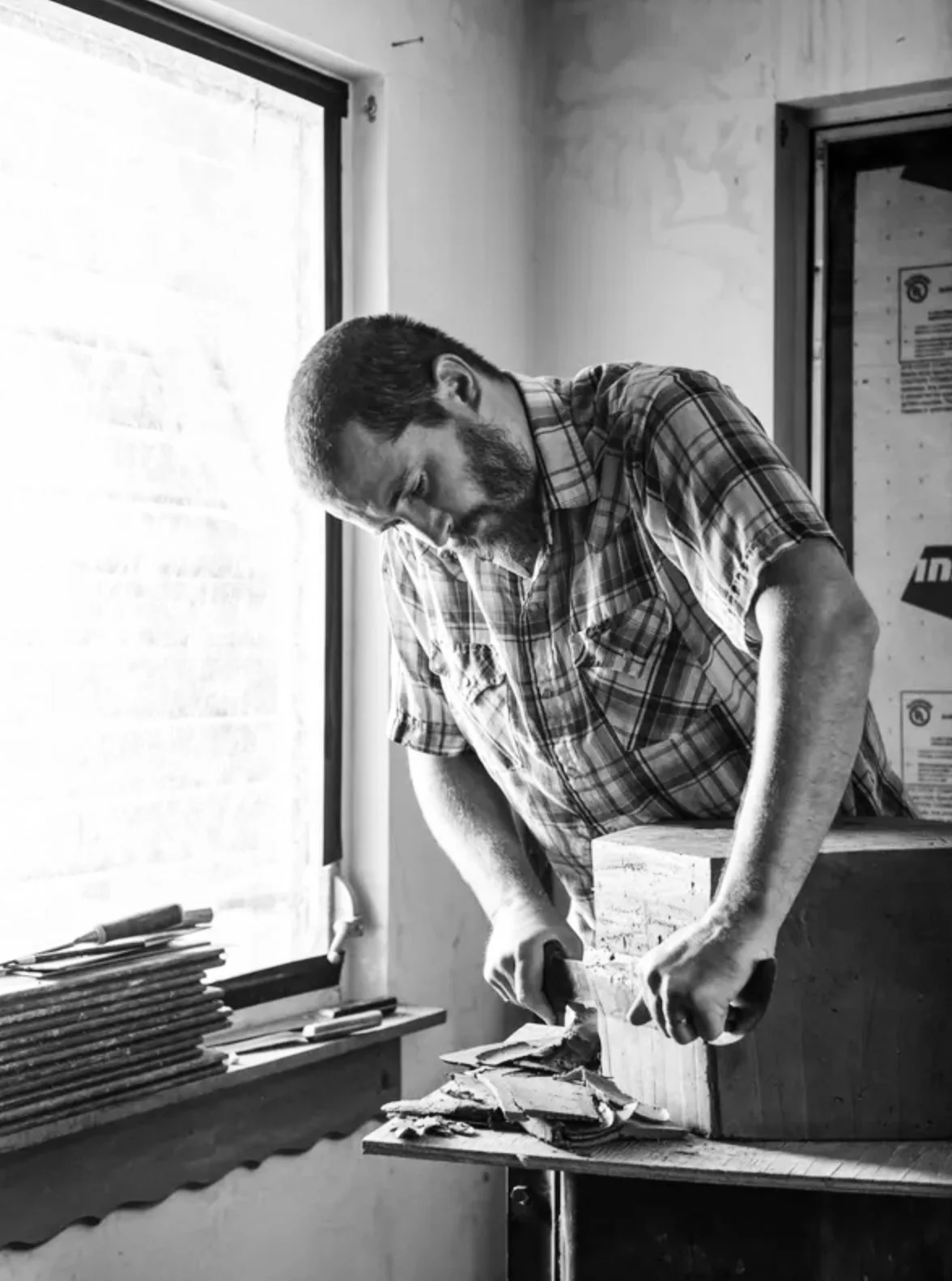 Black and white photo of a man in a plaid shirt woodworking by a window.