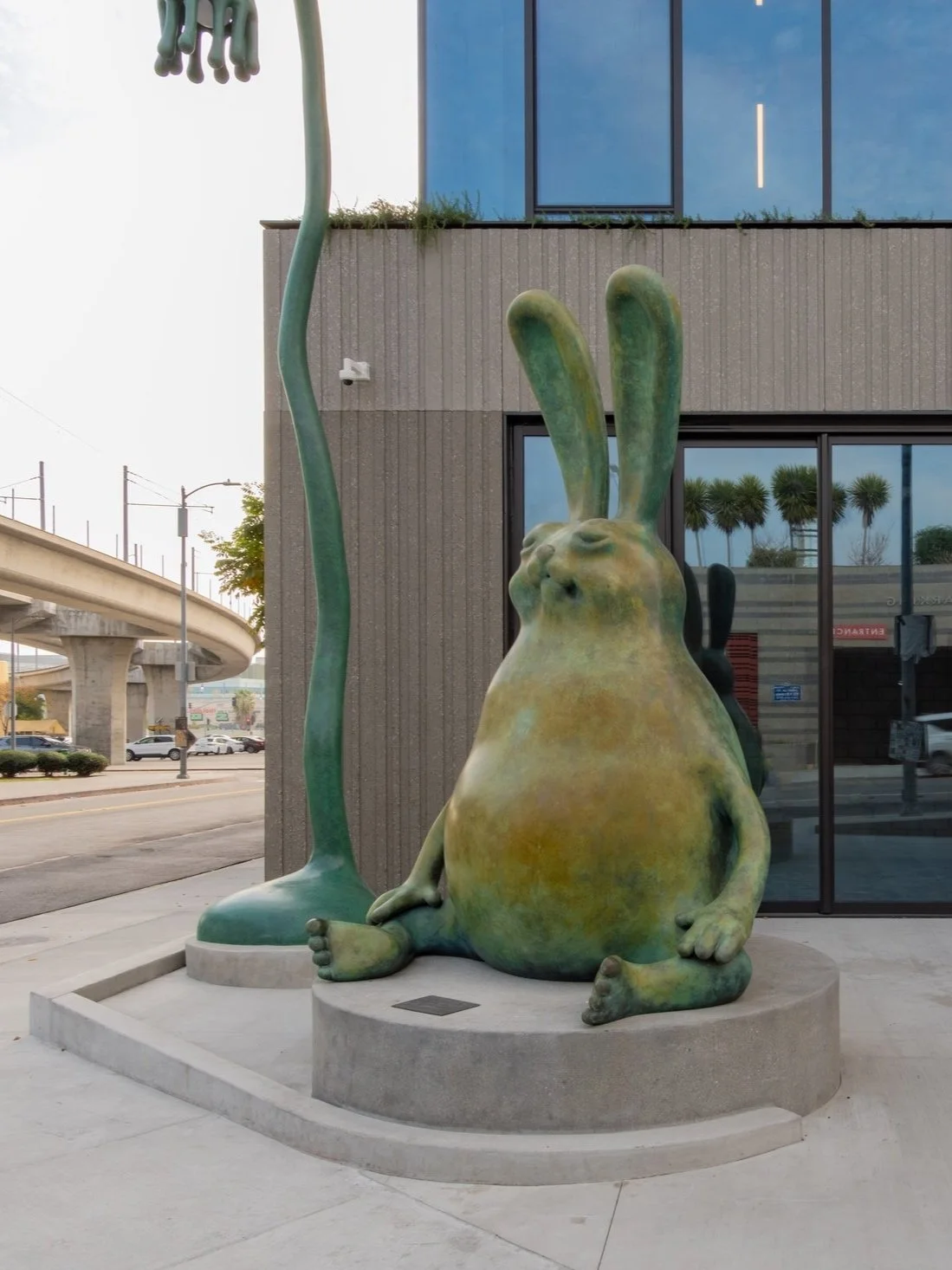 A large sculpture of a green rabbit with an abstract rounded body and long ears, sitting on a circular pedestal outside a modern building, with a tall, thin sculpture of a hand reaching down from above.