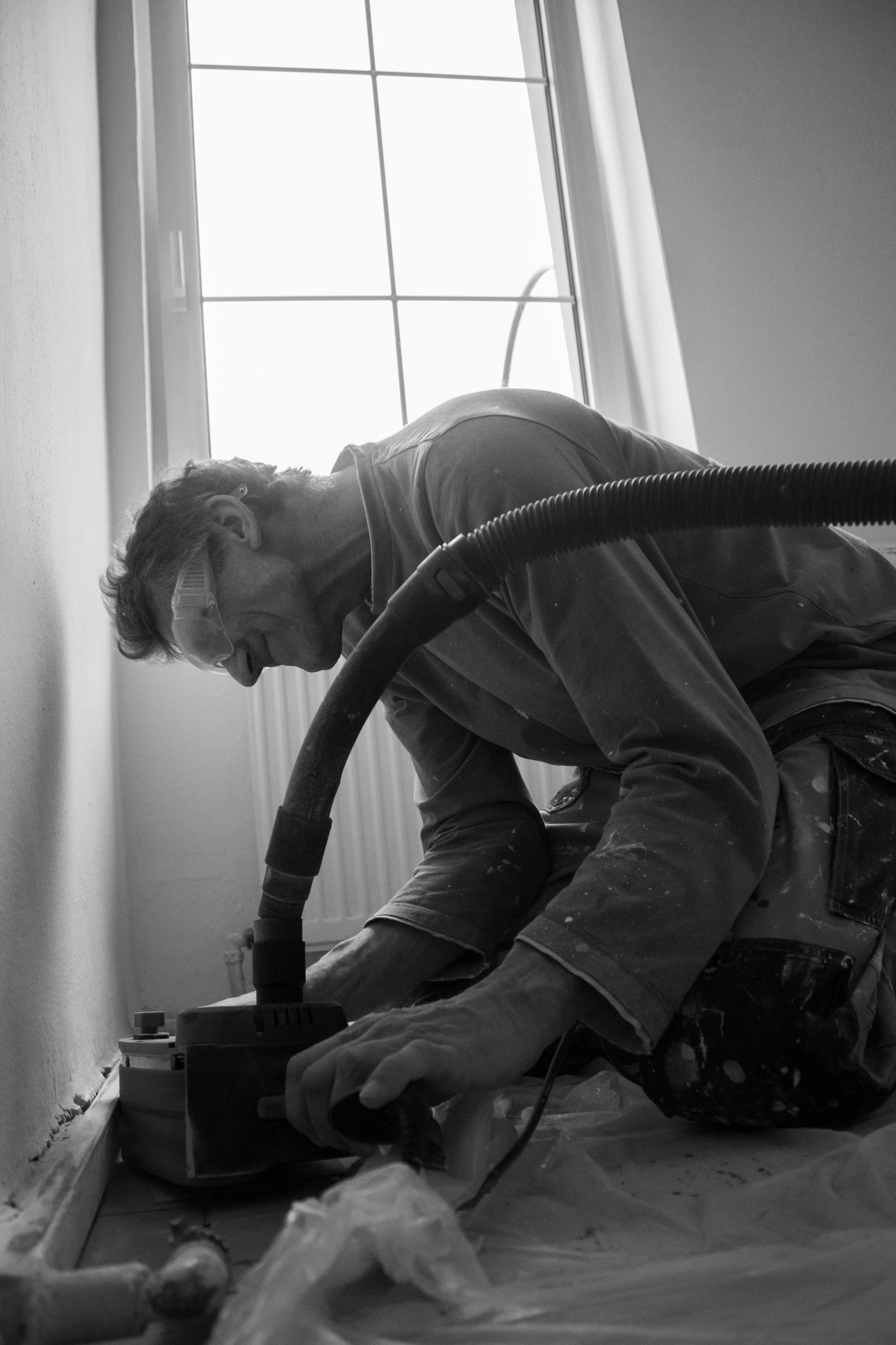 A man working on a home improvement project, using a power sander or drill on the floor near a wall and window, wearing safety goggles.