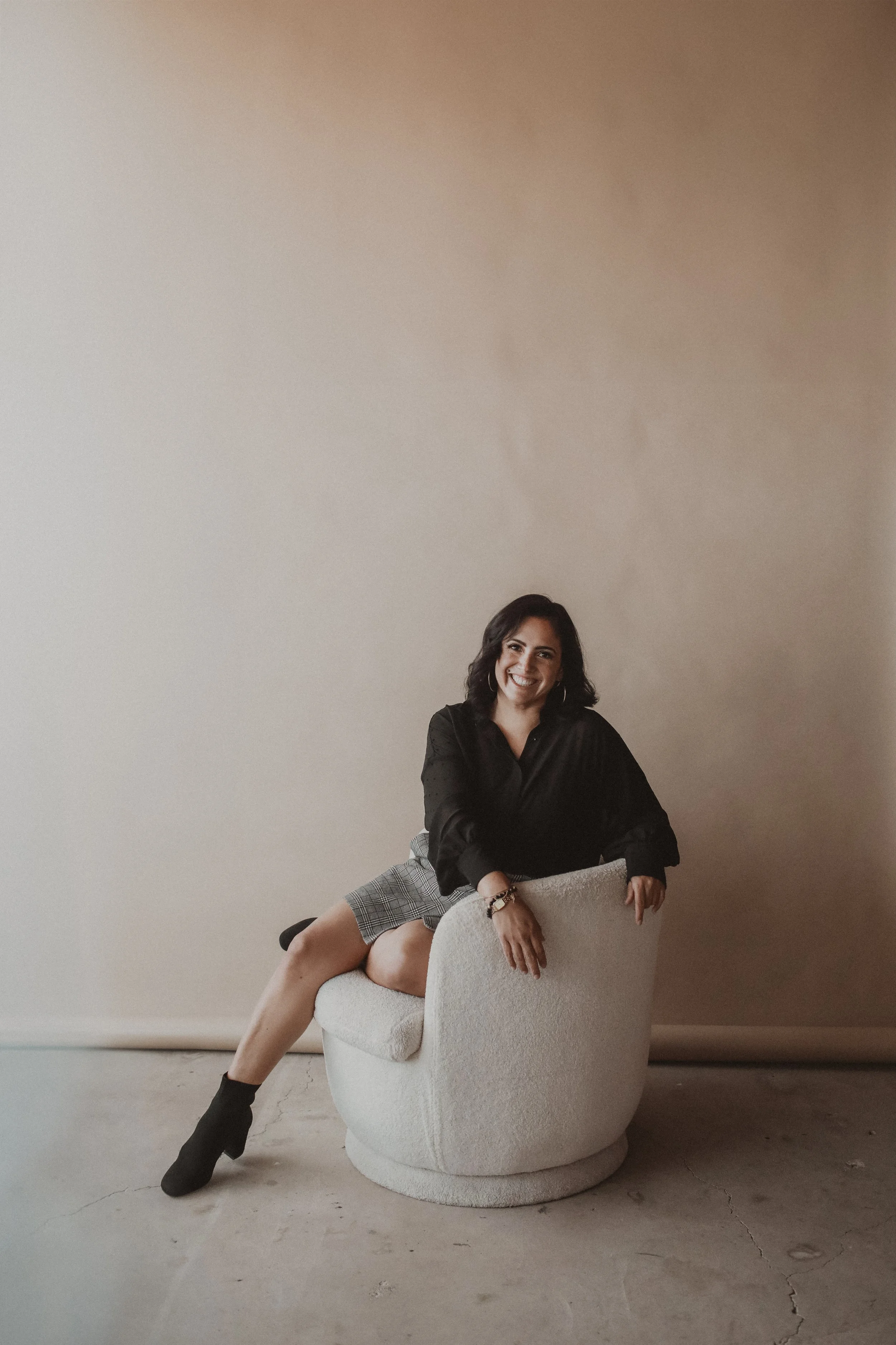 A woman sitting on a round, modern, white upholstered chair, smiling at the camera, against a plain beige wall.