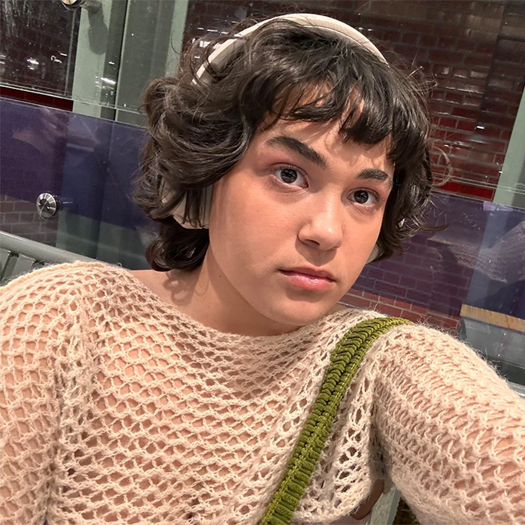 A woman with short, curly brown hair and wearing a pink headband. She has on a pink, open-knit sweater and is holding a green strap. She is looking at the camera with a neutral expression, in an indoor setting with glass and brick walls in the background.