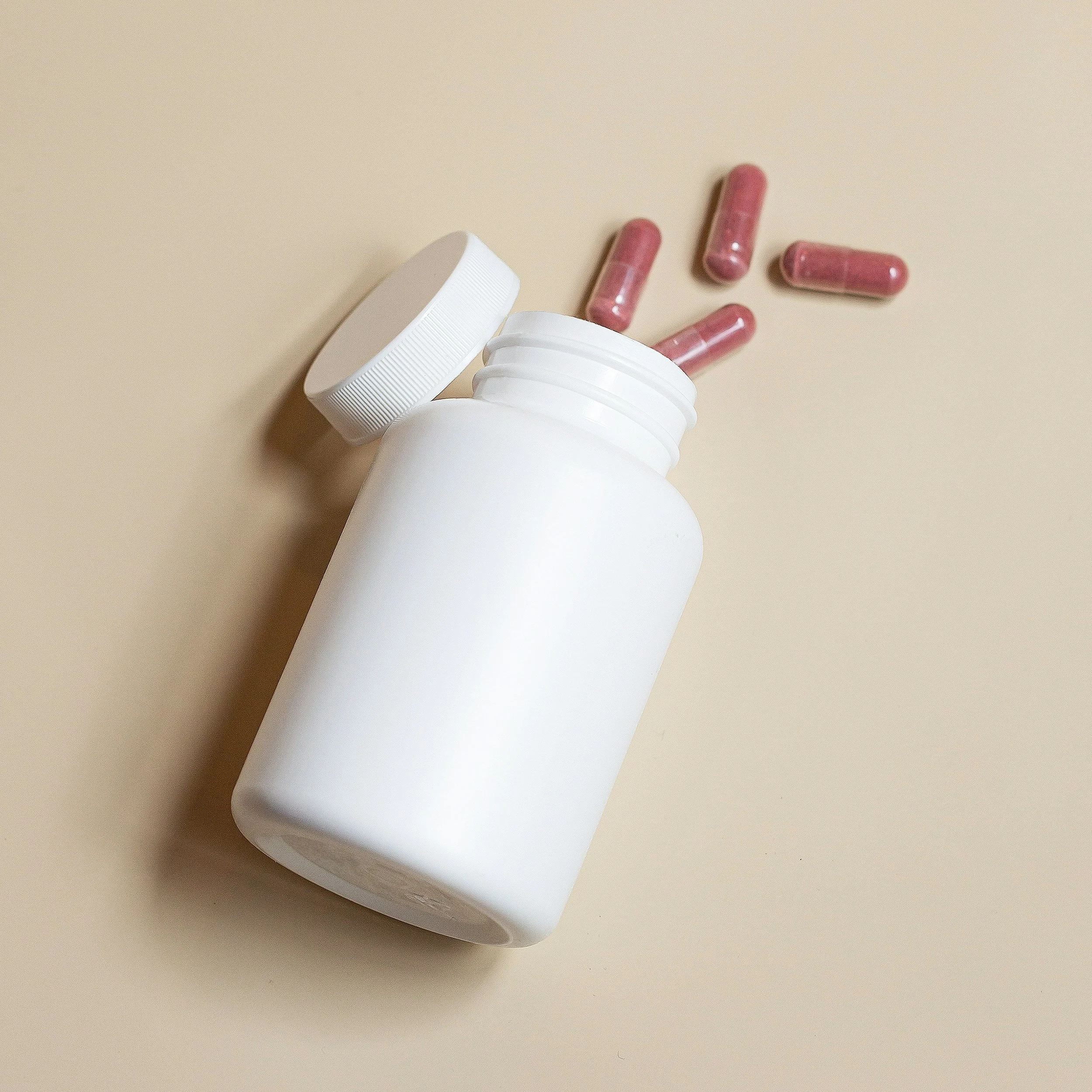 Lege witte pillenpot met roze capsules naast op een beige achtergrond.