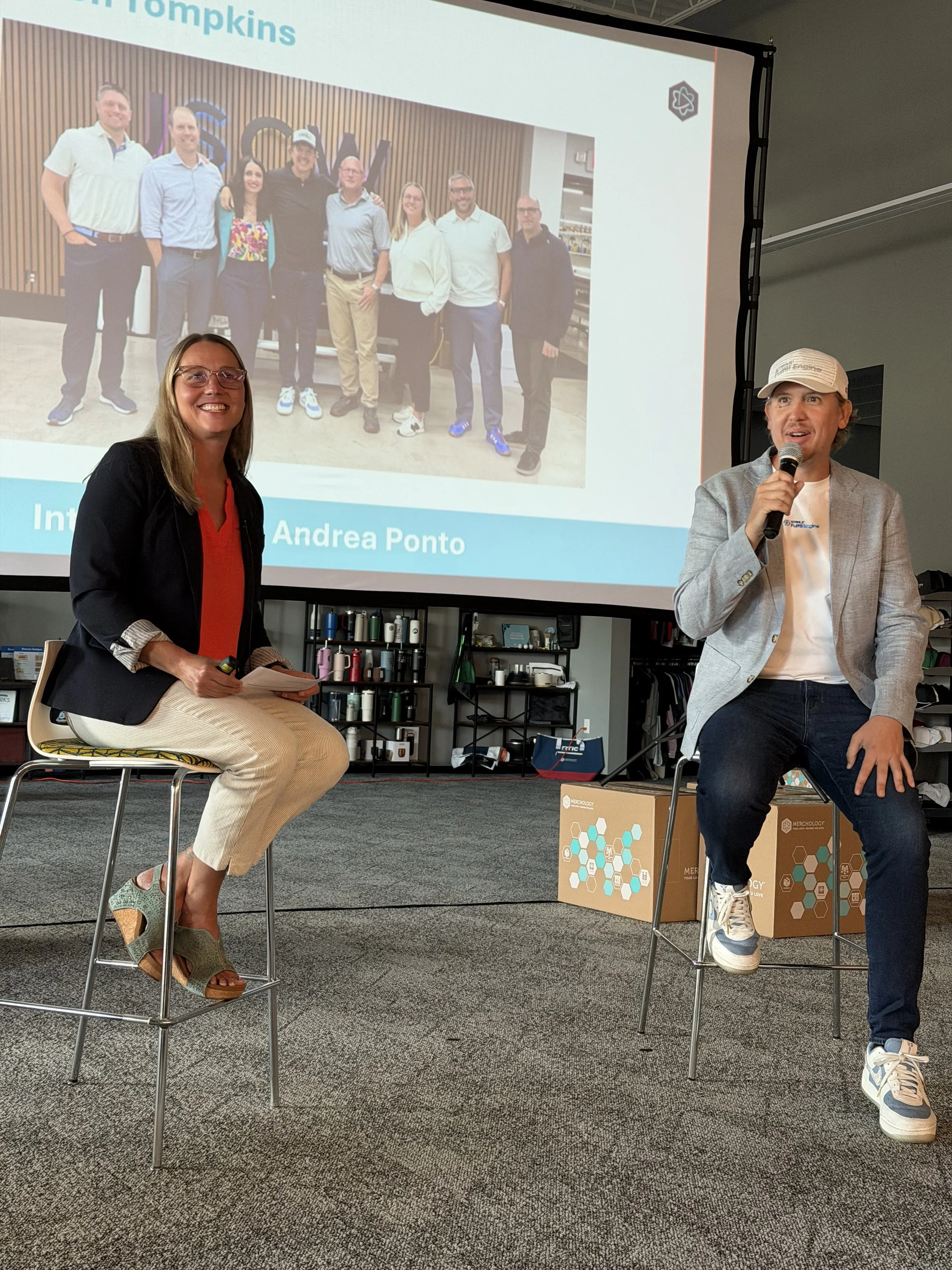 Two people sitting on high stools in front of a presentation screen, with a group photo projected on it. The woman on the left is smiling, wearing a black blazer and beige pants, holding a notepad. The man on the right is also smiling, dressed in a light gray blazer, white t-shirt, jeans, and sneakers, holding a microphone. The background includes shelving with various items and boxes on the floor.