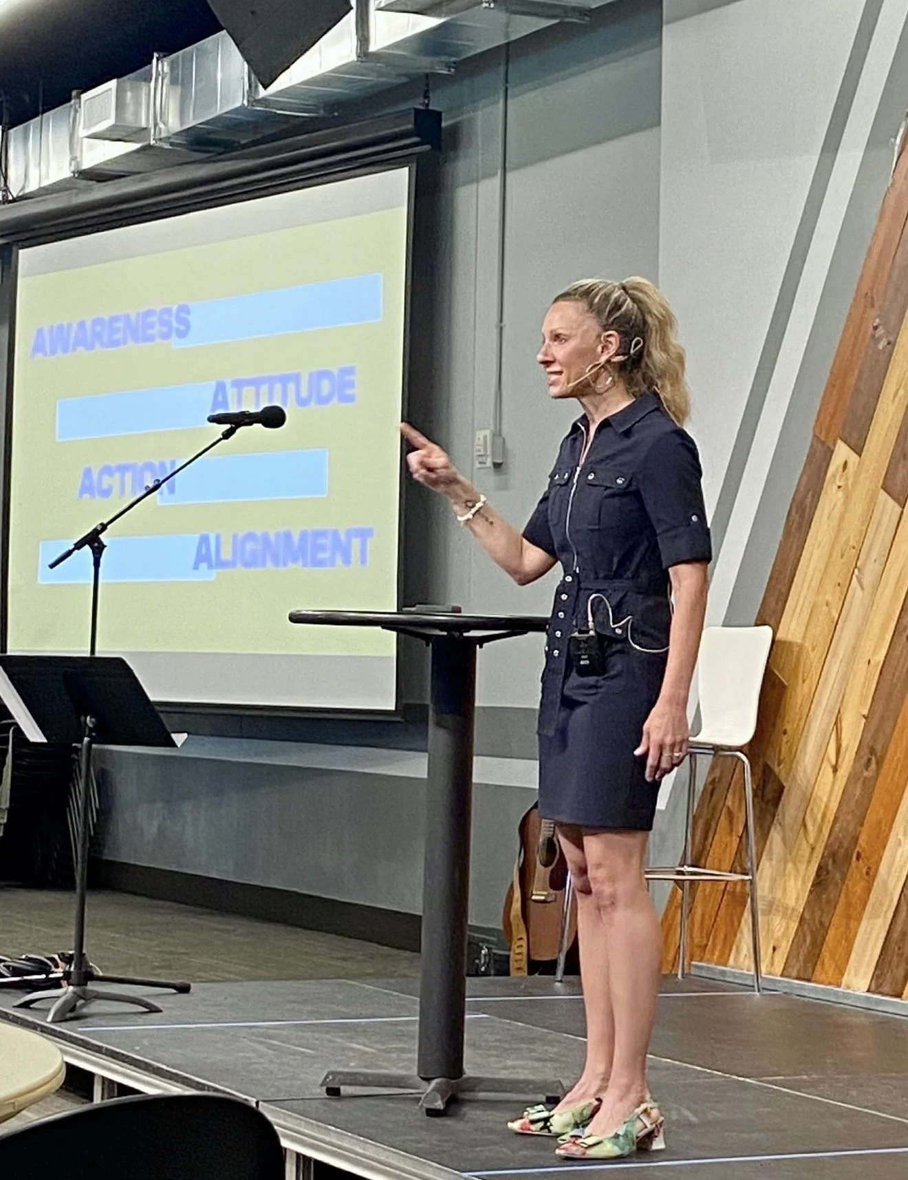 A woman in a black dress, wearing colorful high heels, is giving a presentation in front of a screen displaying words: awareness, attitude, action, and alignment. She appears to be speaking passionately.