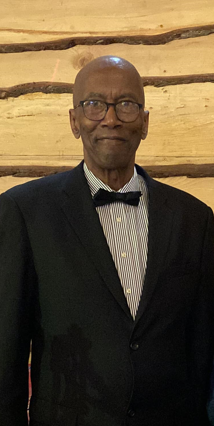 Butch Hill, Board Member of the Charleston, WV Blues Society, dressed in a tuxedo with a striped dress shirt and black bow tie, wearing glasses, standing against a wooden log wall.