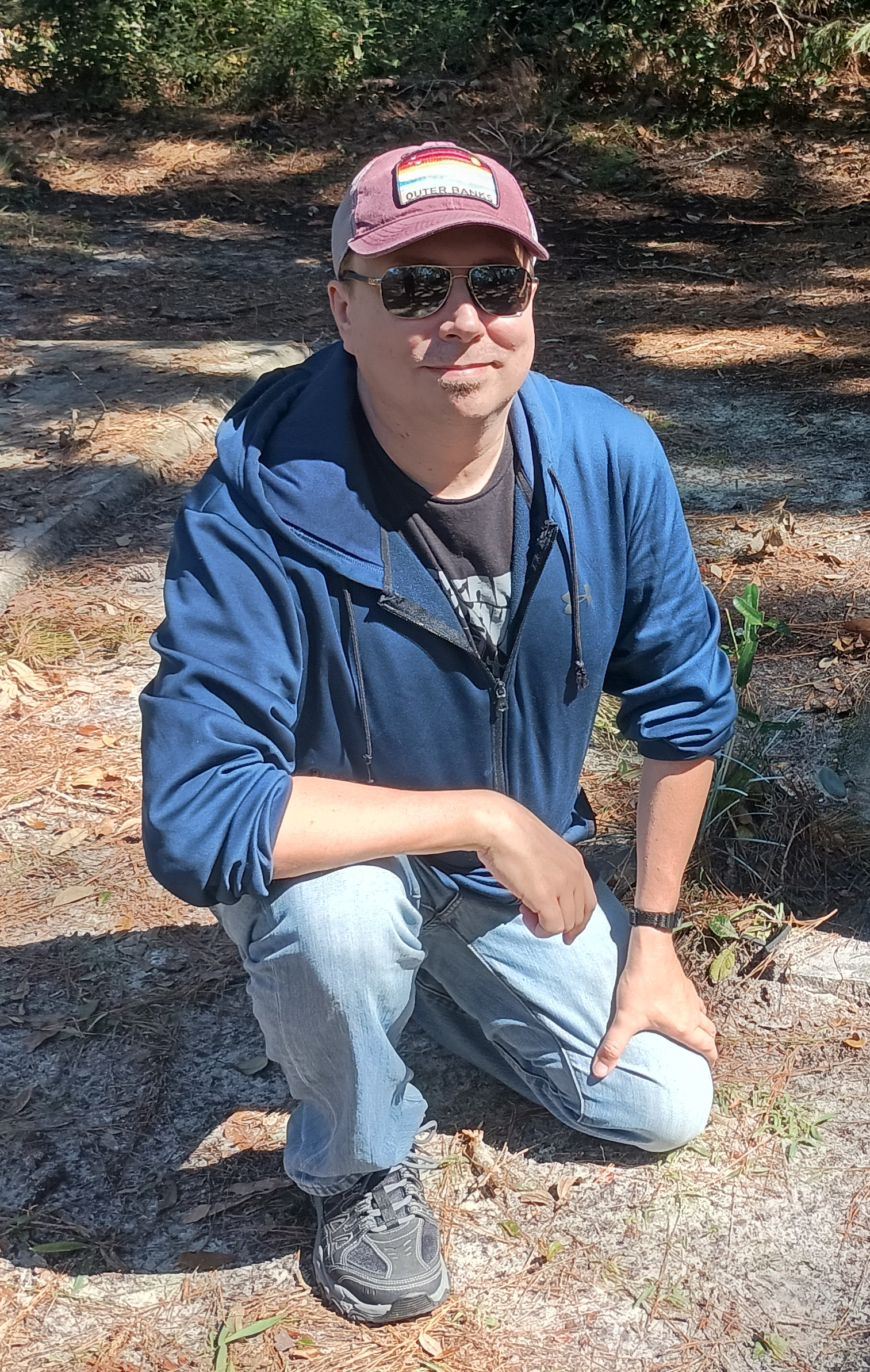 Jason Riffle, Board Member of the Charleston, WV Blues Society, at the Union Cemetery in Saint Simons Island, Georgia, squatting on the ground, wearing sunglasses, a maroon cap with a rainbow patch, a blue jacket, light jeans, and hiking shoes.