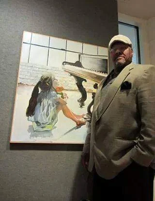 Newman Jackson, Board Member of the Charleston, WV Blues Society, in a beige suit and baseball cap standing in front of a painting of a girl playing a piano.