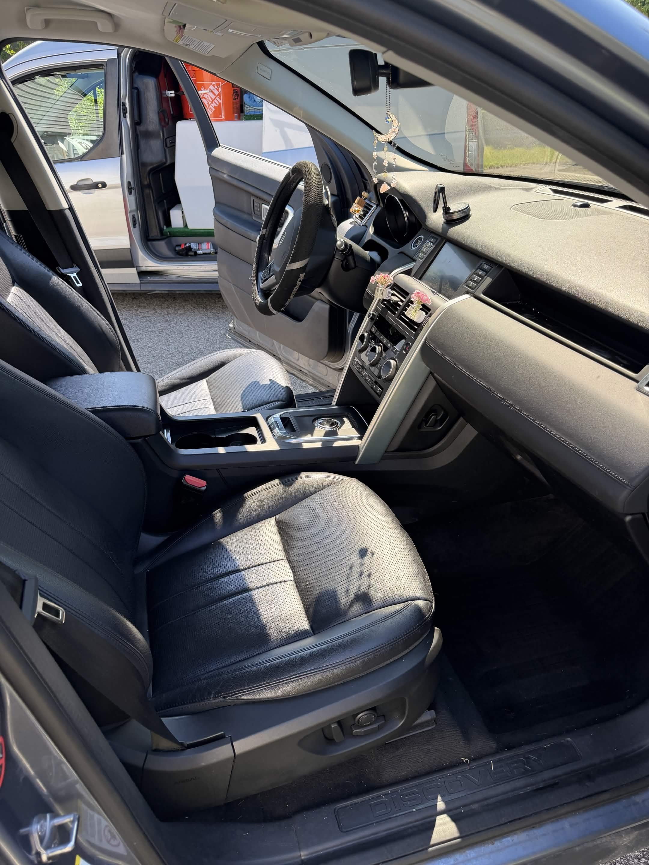 View of the interior of a Nissan vehicle, showing the front passenger seat and dashboard. The dashboard is decorated with hanging jewelry and small flower arrangements. Outside the open car door, a pickup truck is visible, with various boxes and item