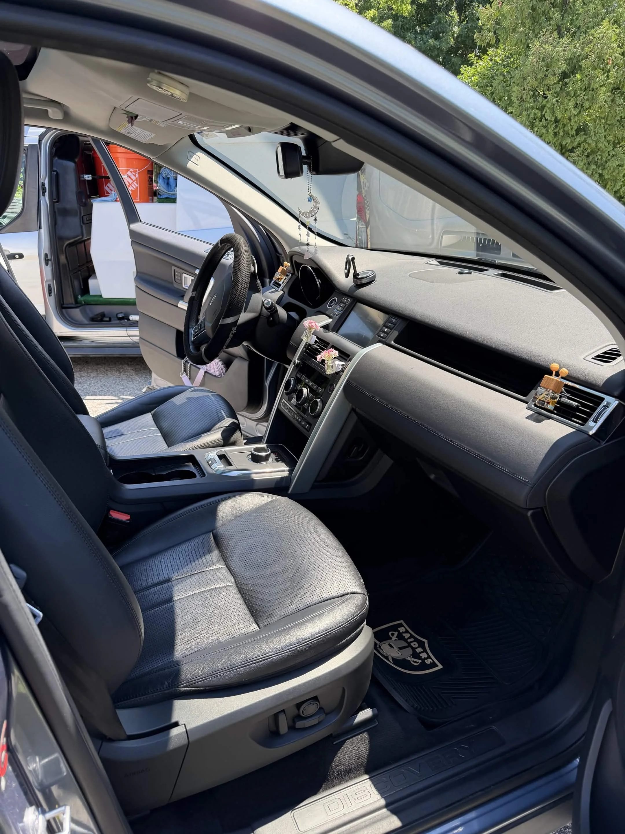 Inside a vehicle showing a black and gray seat, dashboard with various controls, and a hanging charm with a crescent moon shape. Part of the car's interior and a bright outdoor scene with green trees are visible.