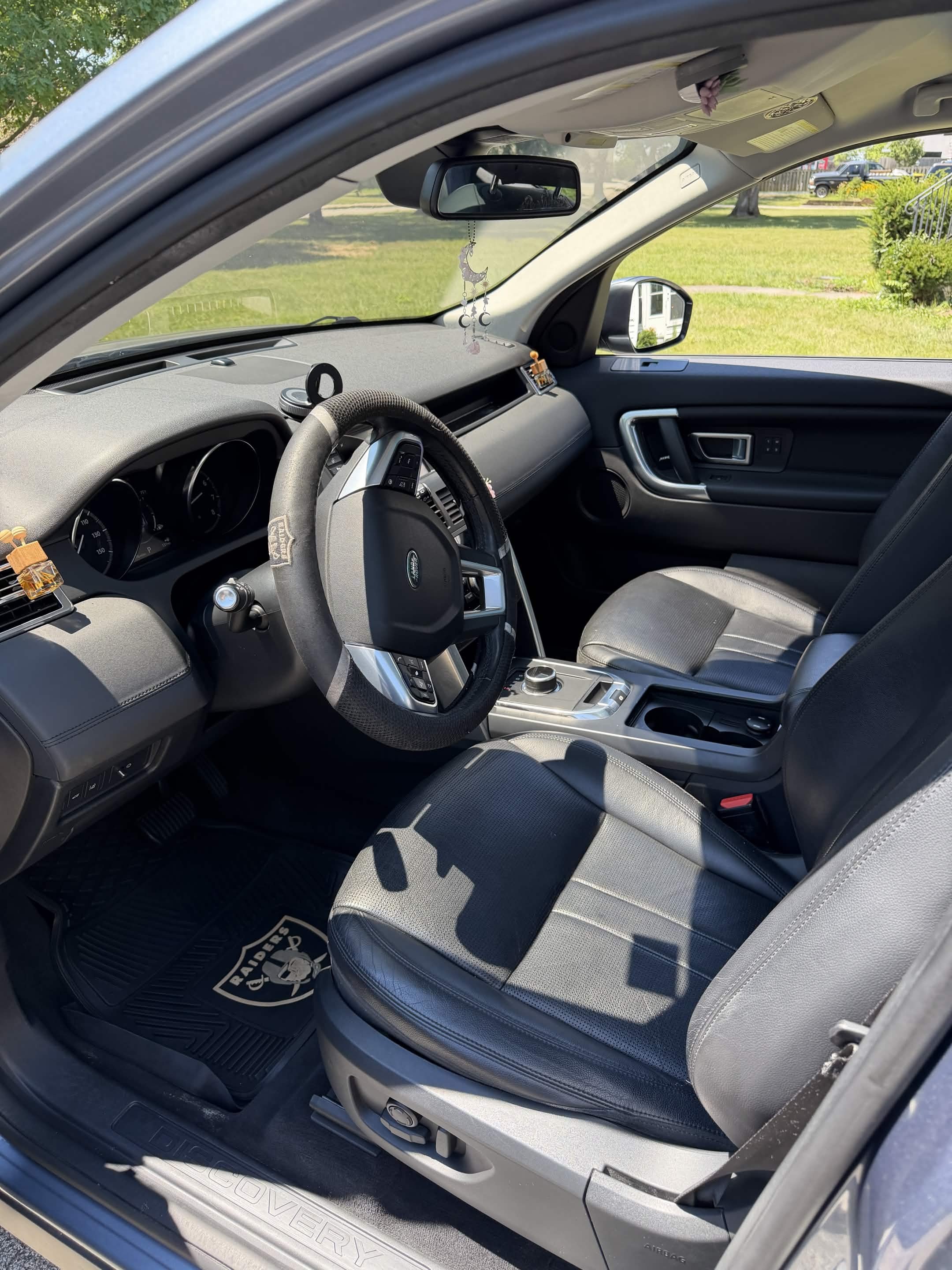 Interior of a gray Land Rover vehicle showing the steering wheel, dashboard, front passenger seat, and side window with a view of a grassy yard and trees outside.