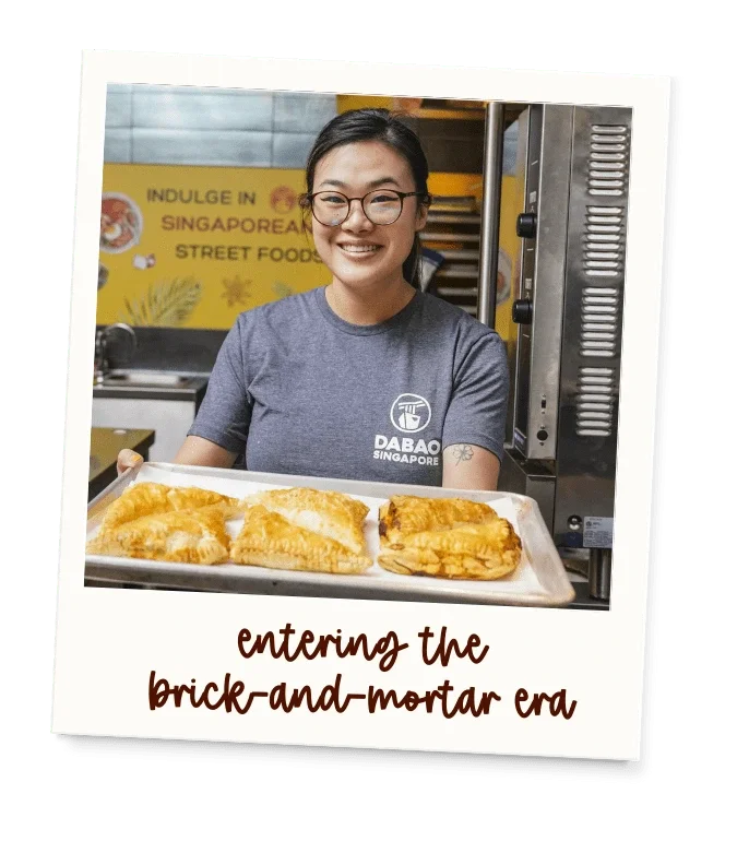 A woman wearing glasses and a gray Daba Singapore T-shirt stands behind a tray of twenty-four fried snacks, likely pasties, in a street food stall with a yellow sign in Singapore.