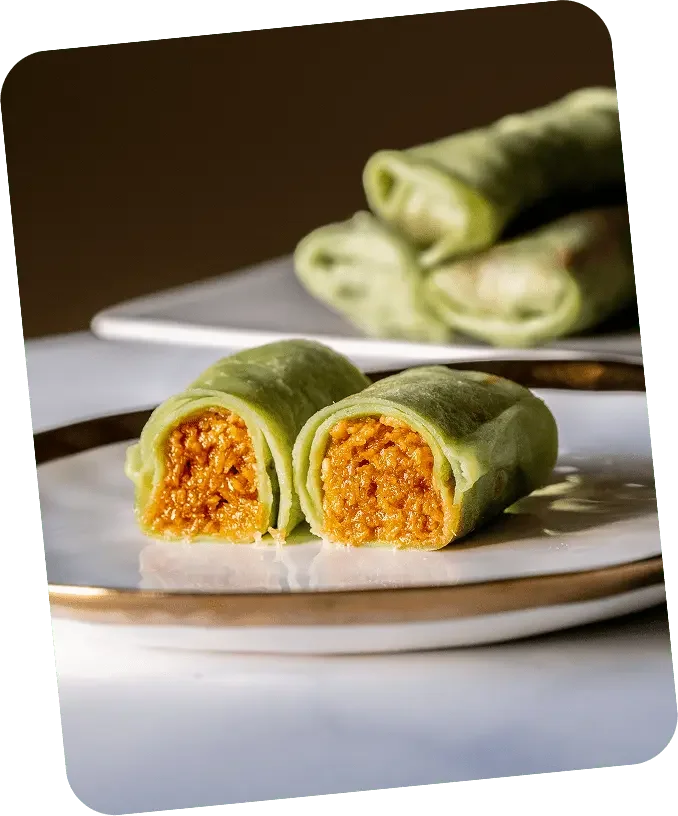 Close-up of a green wrap sliced in half, revealing an orange filling, on a white plate with a gold rim. In the background, more wraps are stacked on a white plate.