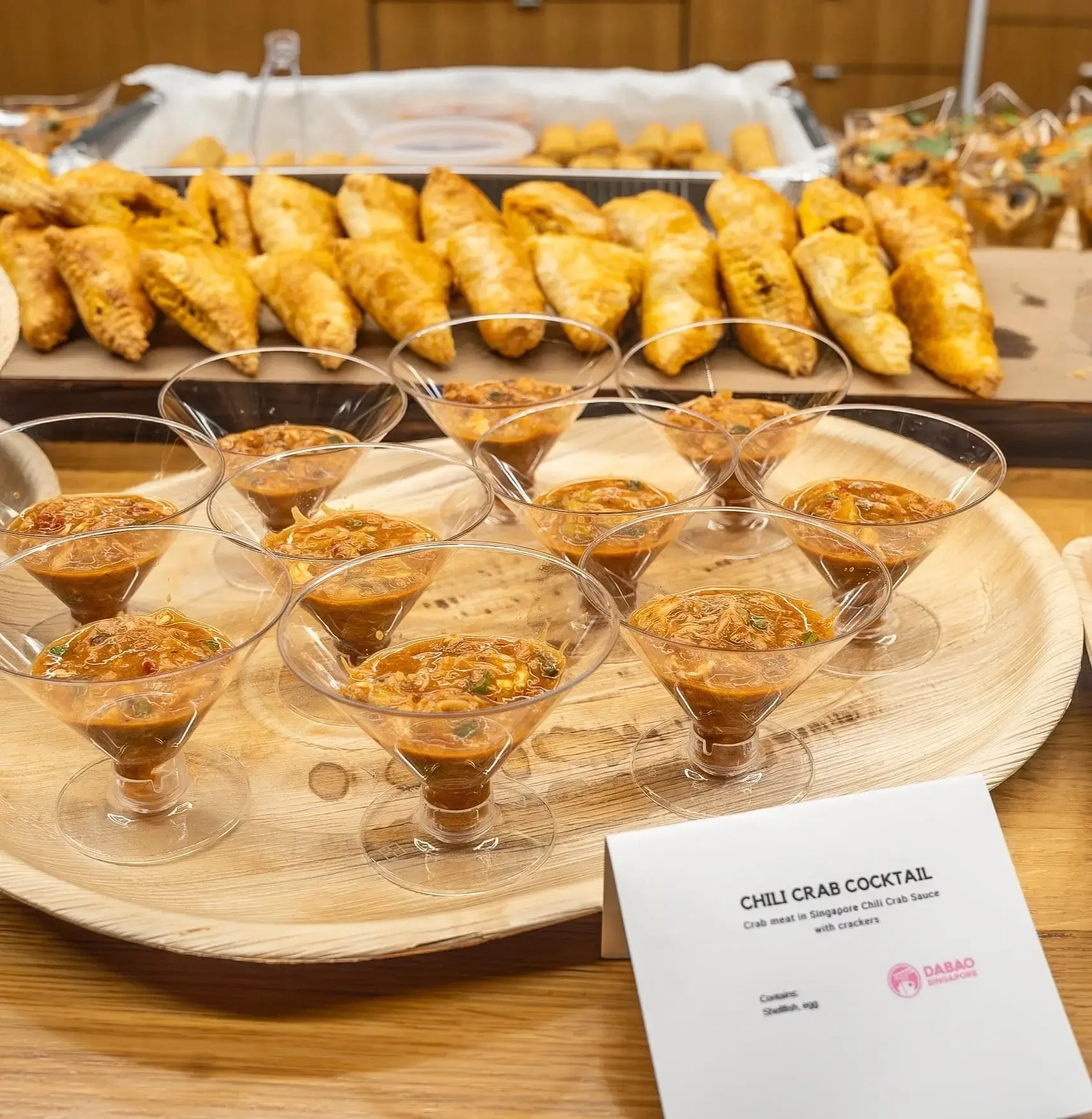 Small glasses of chili crab cocktail with crab meat in Singapore chili crab sauce, served with crackers on a wooden tray. In the background, there are fried pastries or spring rolls arranged on a wooden tray.