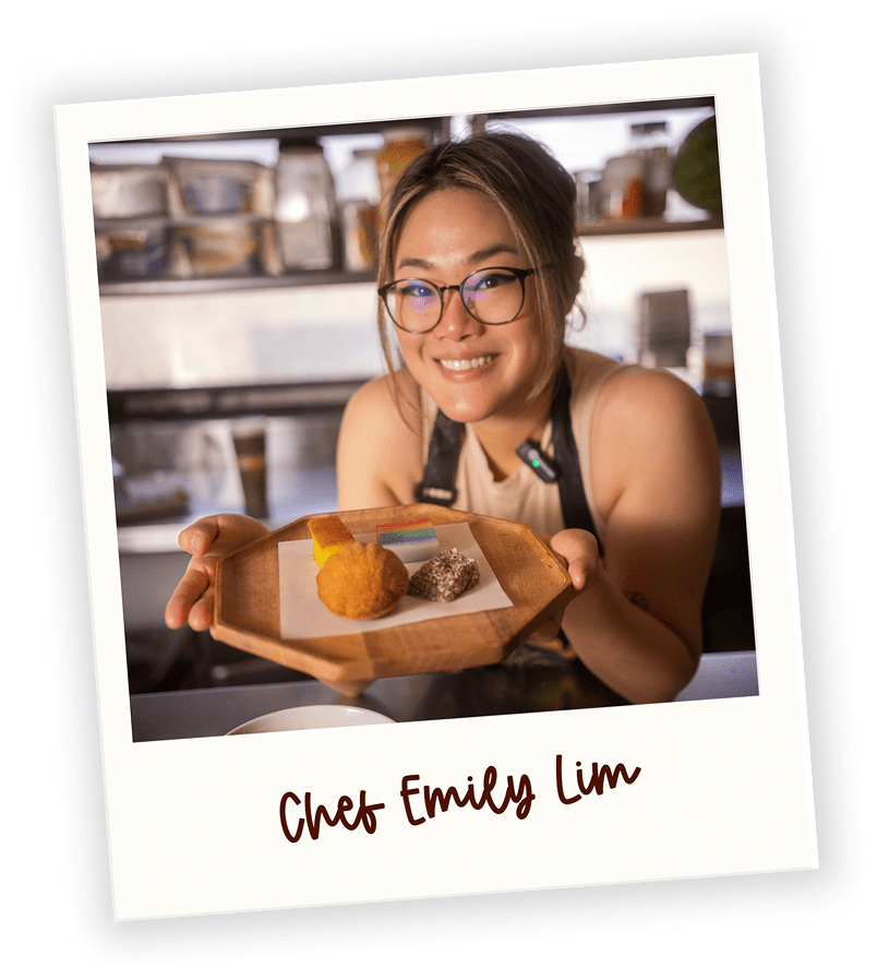 A smiling woman with glasses and a chef's apron presenting a wooden tray with three food items, in a kitchen setting.