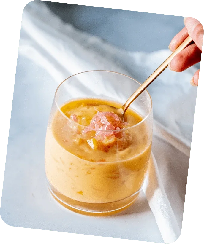 A glass of yellowish dessert with chunks of pink ham and a spoon, held by a hand, on a white surface with a white cloth in the background.