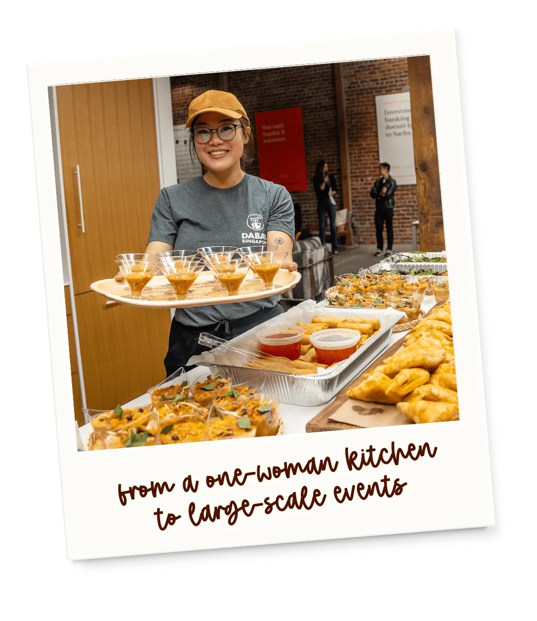 A woman wearing glasses and a yellow cap stands behind a table with various small food dishes and sauces, smiling at the camera at a large social event.