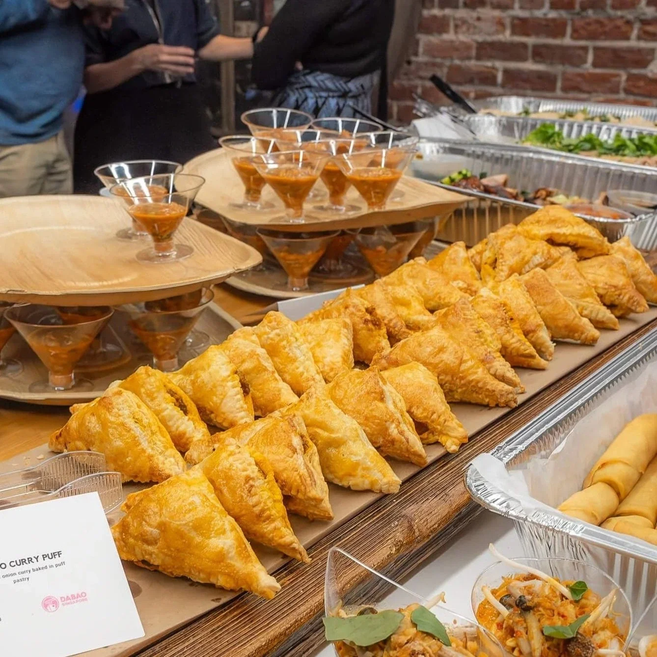 A display of various Asian food items on a buffet table, including golden fried samosas, small glasses of curry or sauce, and other dishes, with people in the background.