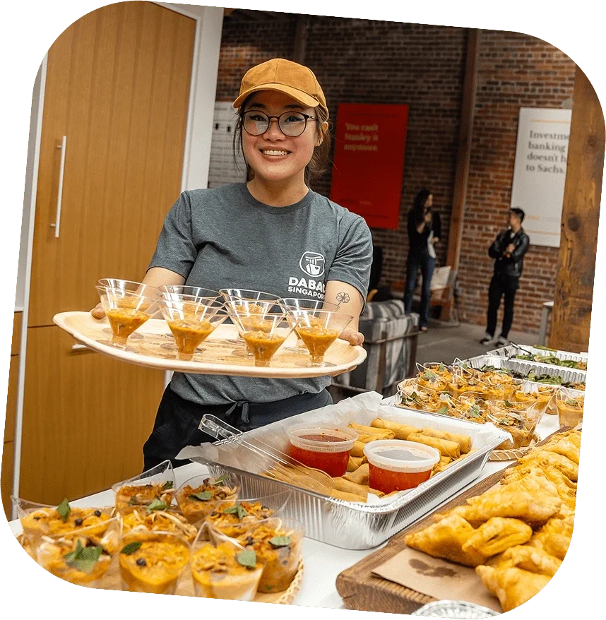 A woman smiling and holding a tray of small glasses filled with a dessert at a buffet table. The table has various snacks and appetizers, including baked items and trays of food, in a room with brick walls and other people in the background.