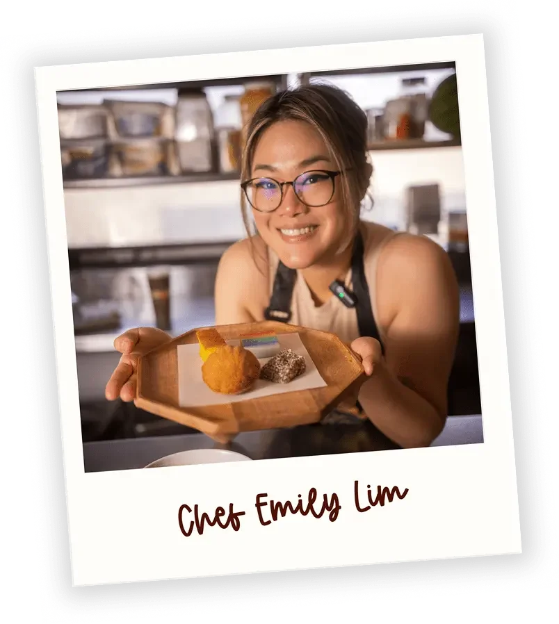 A smiling woman with glasses offers a plate with two treats, a fried item and a powdered dessert, in a kitchen. The photo has a caption reading 'Chef Emily Lim'.