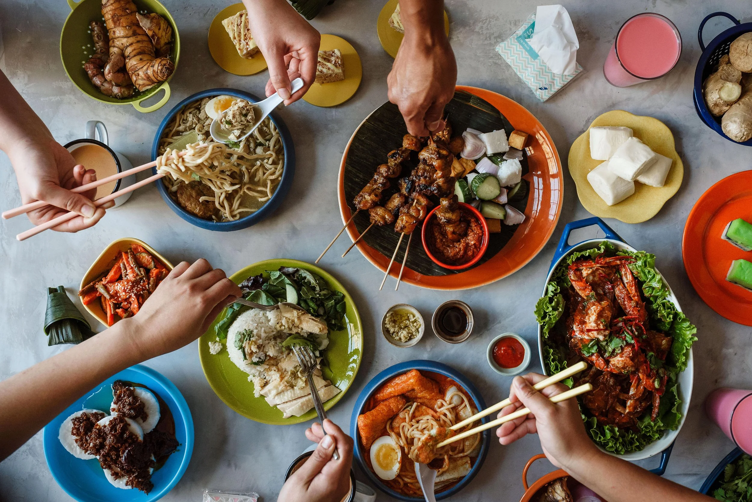 A variety of Asian dishes on a table, including skewers, noodles, rice, vegetables, and desserts, with hands reaching in to serve or eat.