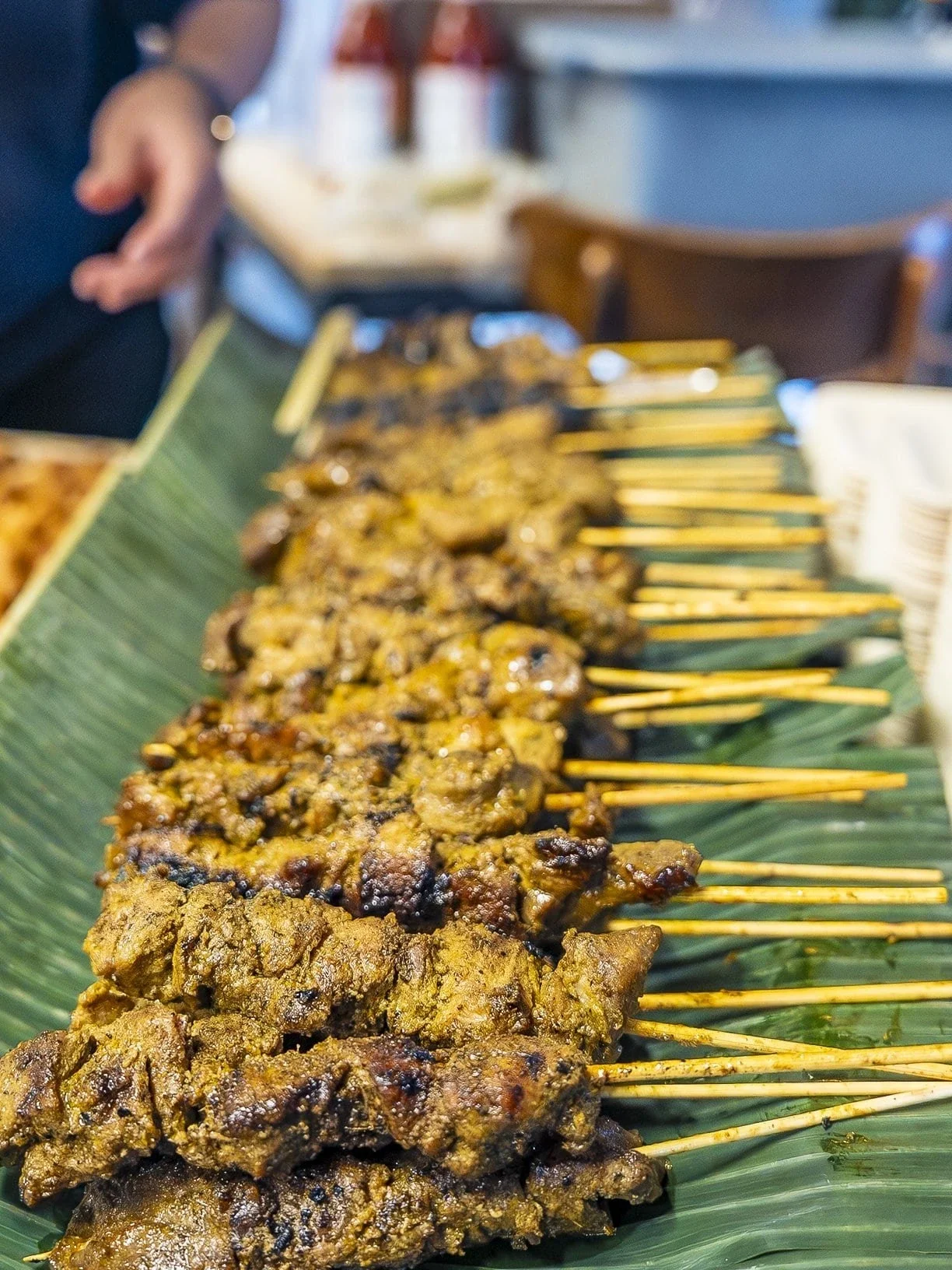Close-up of grilled meat skewers on a banana leaf, with a blurred background of condiments and a person reaching toward the food.