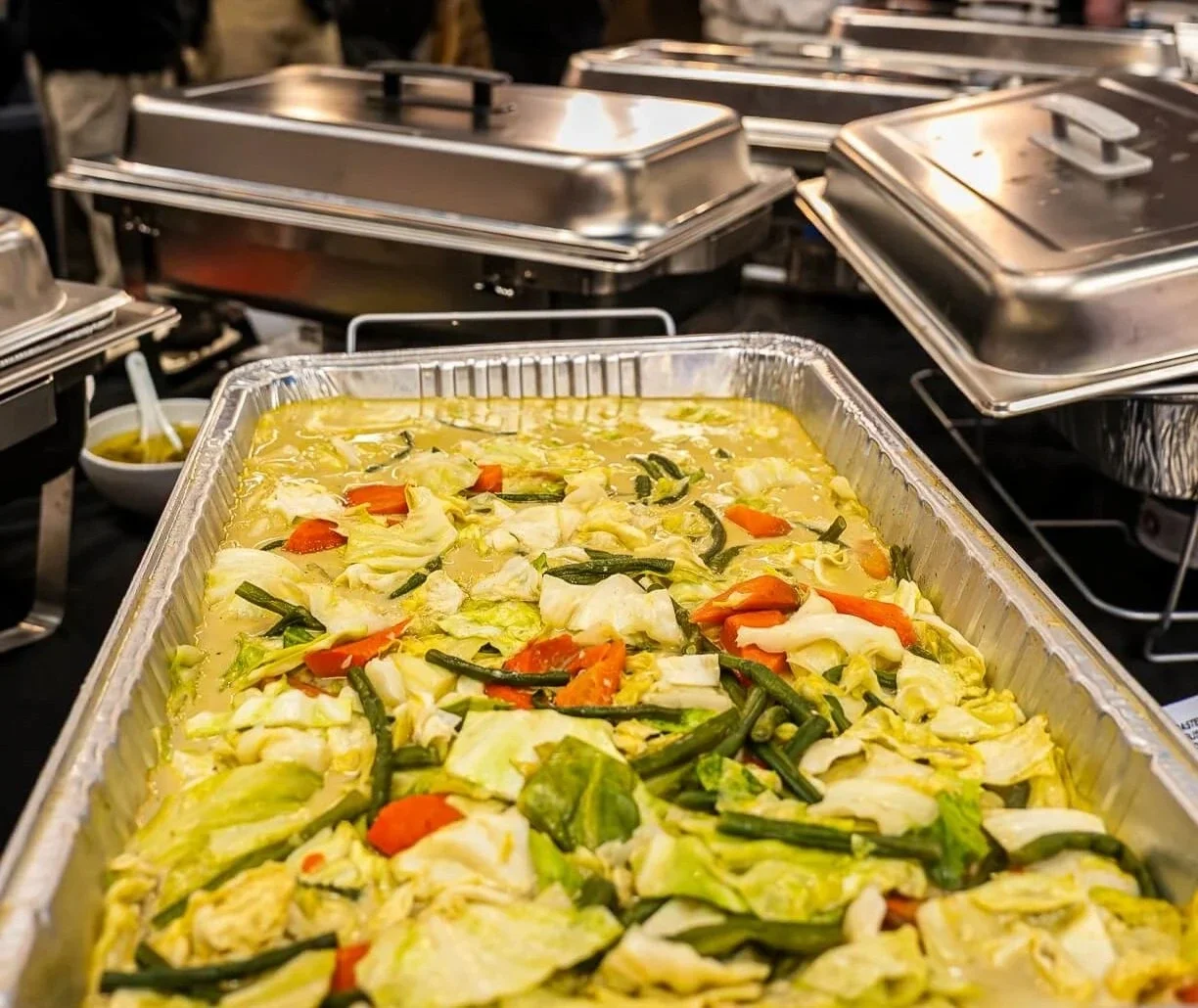 A large tray of cooked vegetables, including cabbage, carrots, and green beans, in a buffet setting with metal chafing dishes in the background.