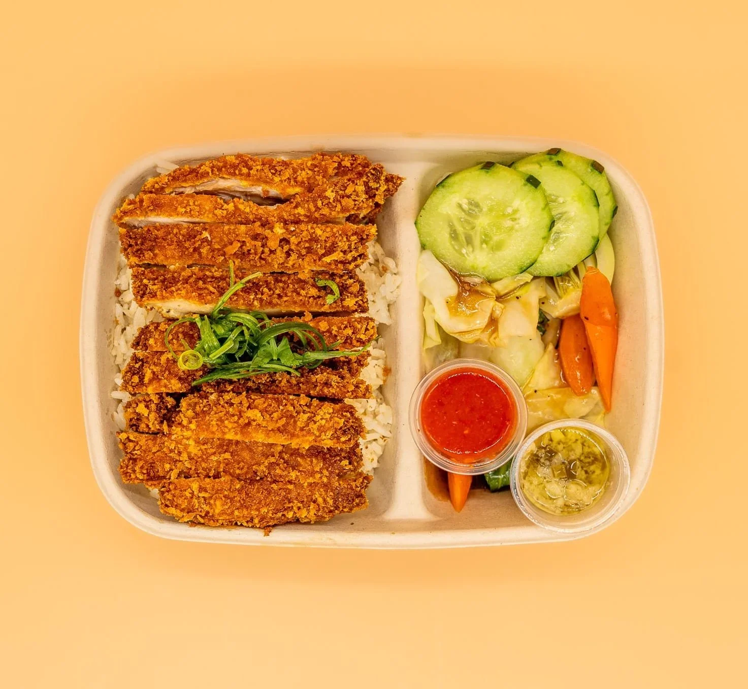 Bento box meal with fried chicken cut into strips on rice, cucumber slices, shredded lettuce, carrots, and two small containers of sauce.